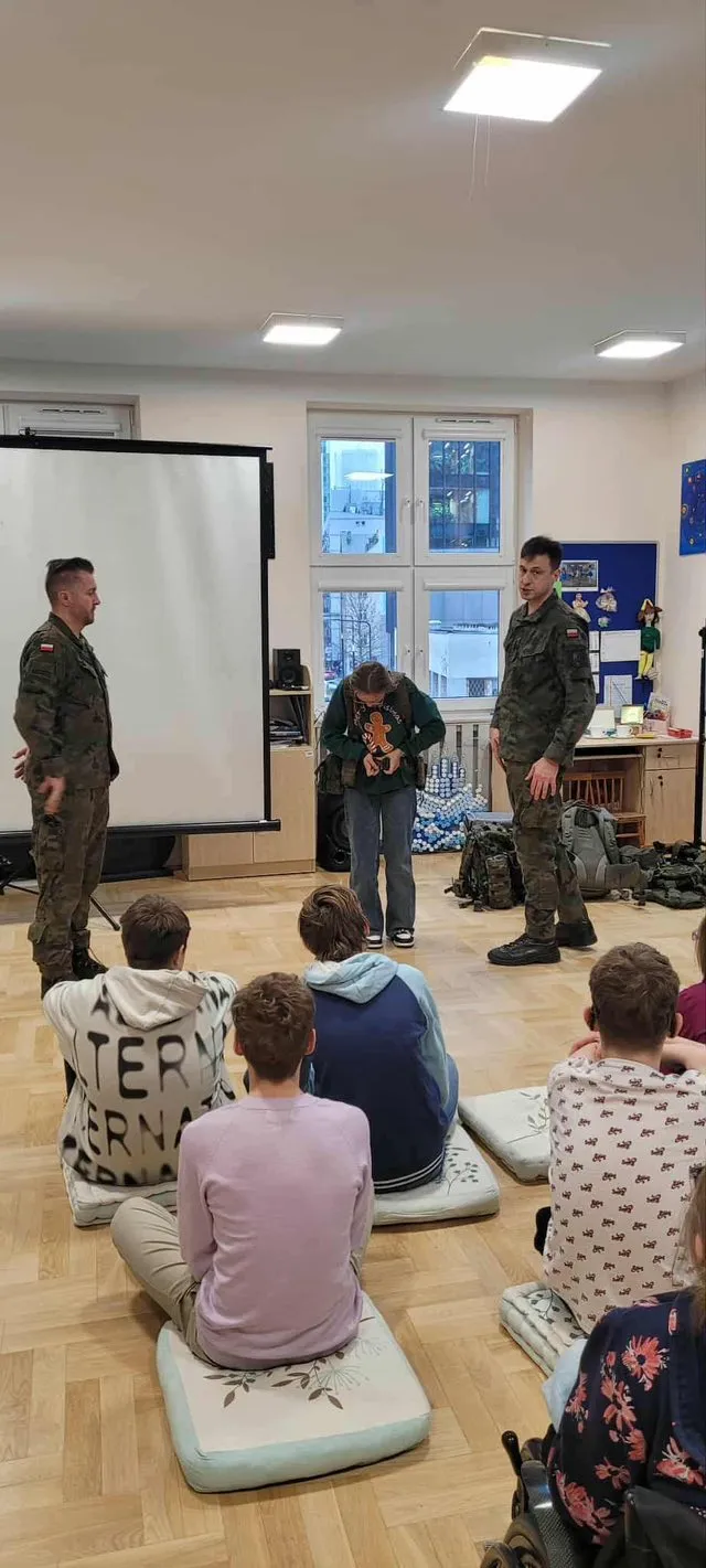 Uczniowie siedzący na podłodze w sali podczas zajęć tematycznych pod kierunkiem  żołnierzy