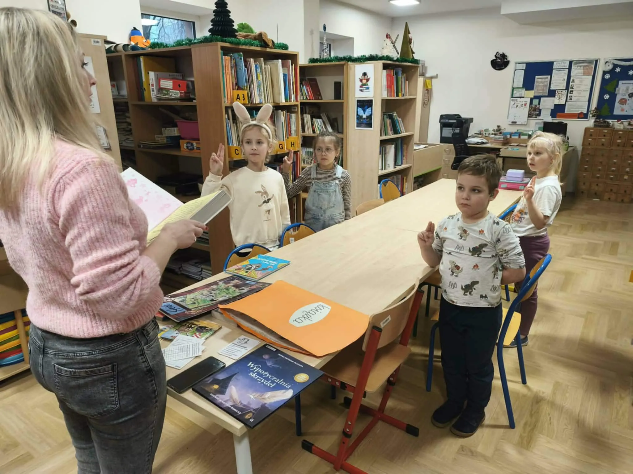 Nauczycielka czyta, dzieci stoją przy stole.