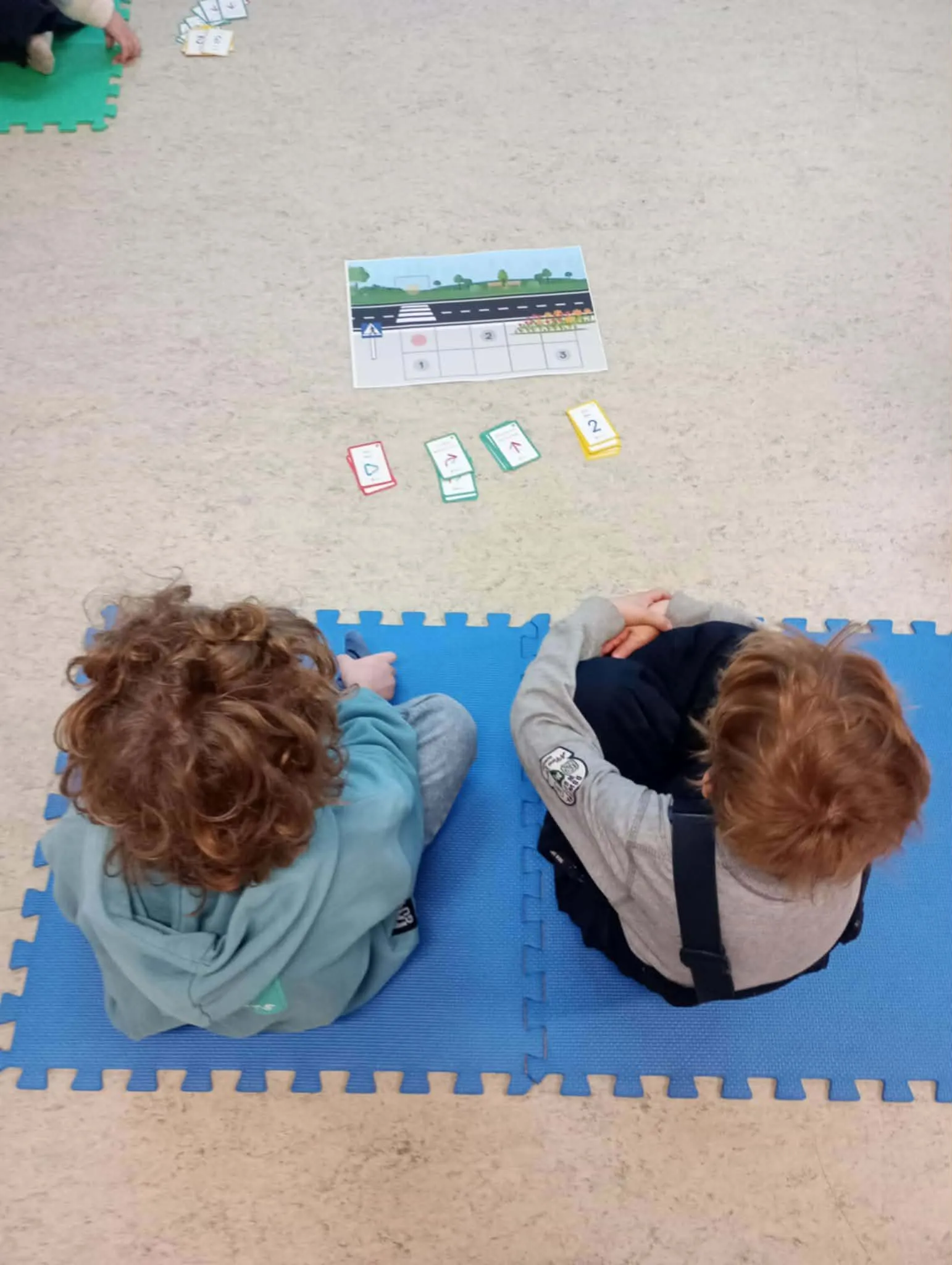 Dwójka dzieci siedzi na niebieskiej macie i obserwuje planszę z przejściem dla pieszych oraz kartami do nauki zasad ruchu drogowego.