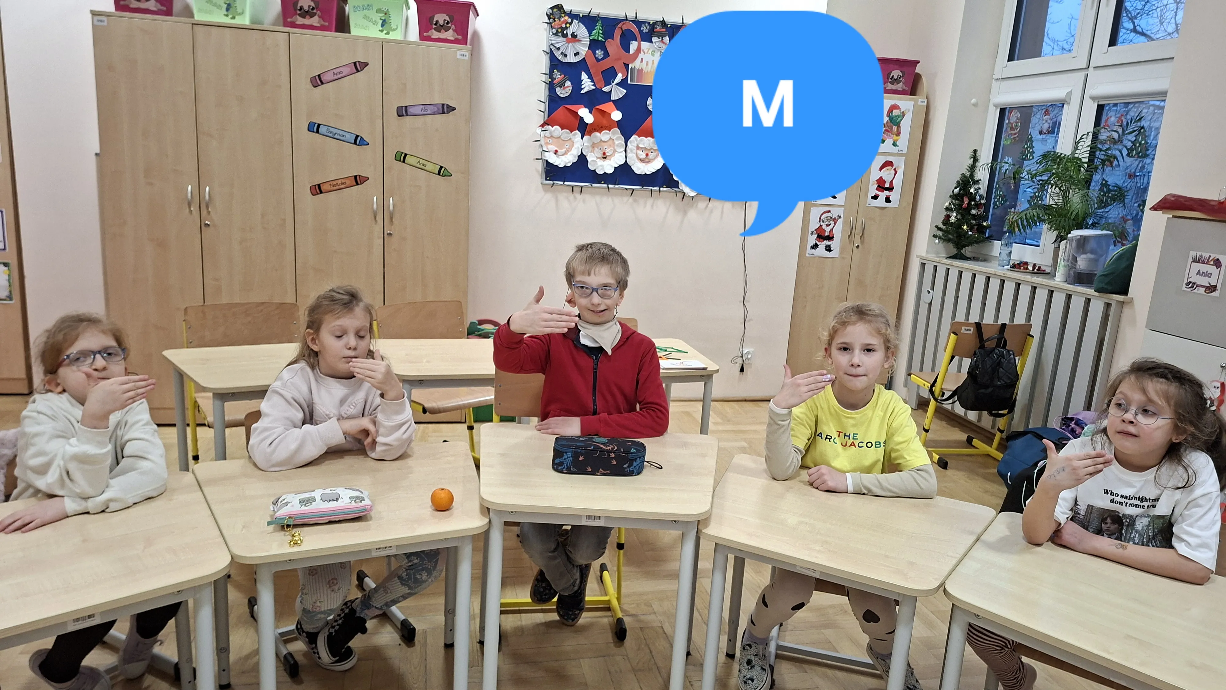 Dzieci siedzą w ławkach i wykonują ustalone gesty dłoni podczas wspólnych ćwiczeń komunikacyjnych w klasie.