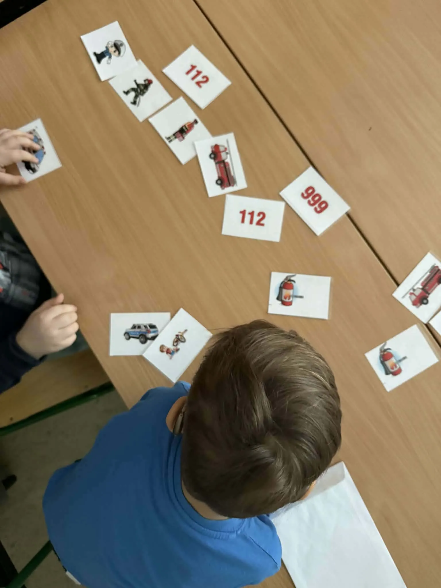 Dzieci przy stoliku układają karty edukacyjne z numerami alarmowymi i ilustracjami służb ratunkowych.