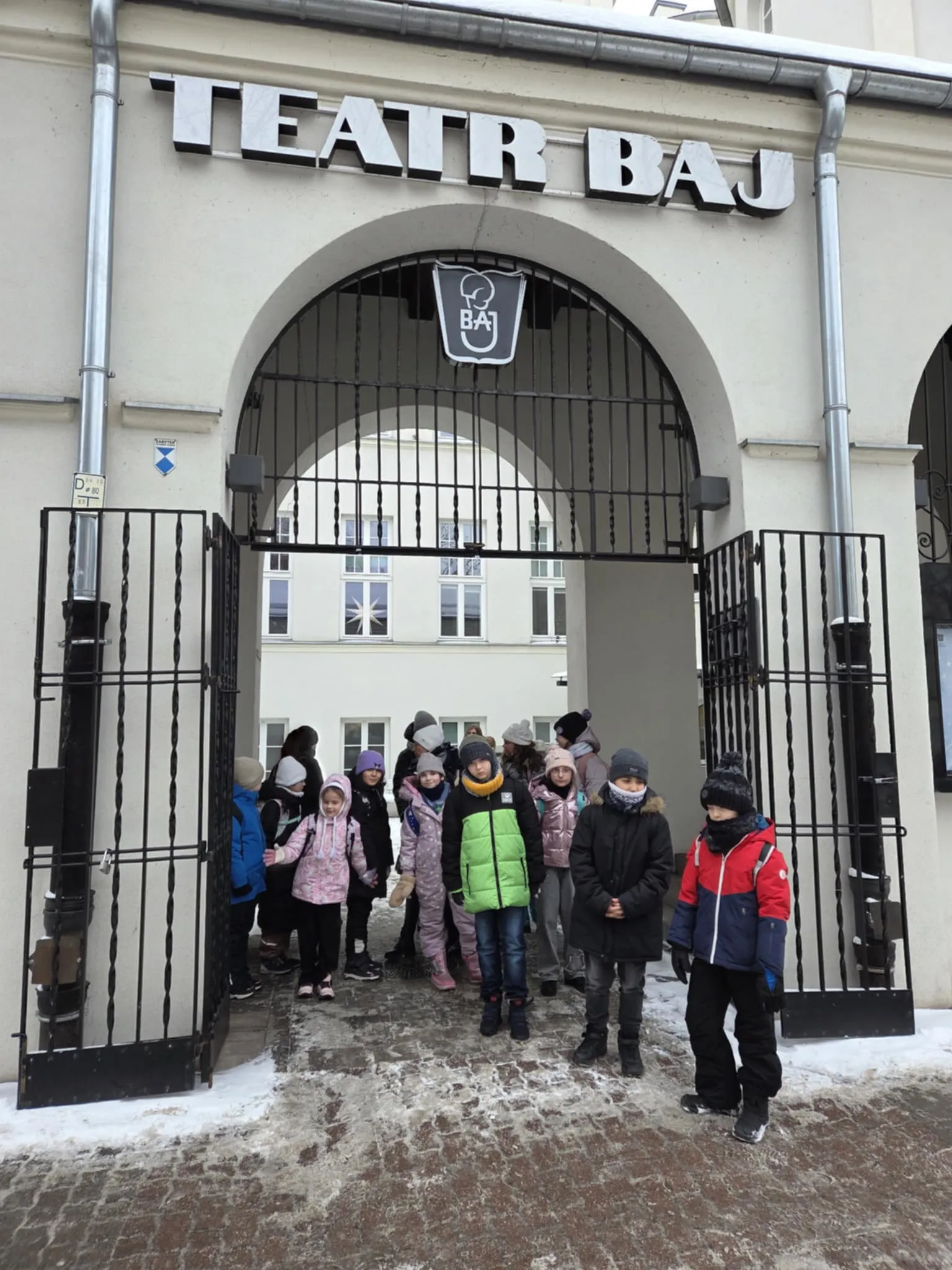 Grupa dzieci ubranych w zimowe kurtki stoi przed wejściem do Teatru Baj, pod charakterystycznym łukiem z napisem teatru.