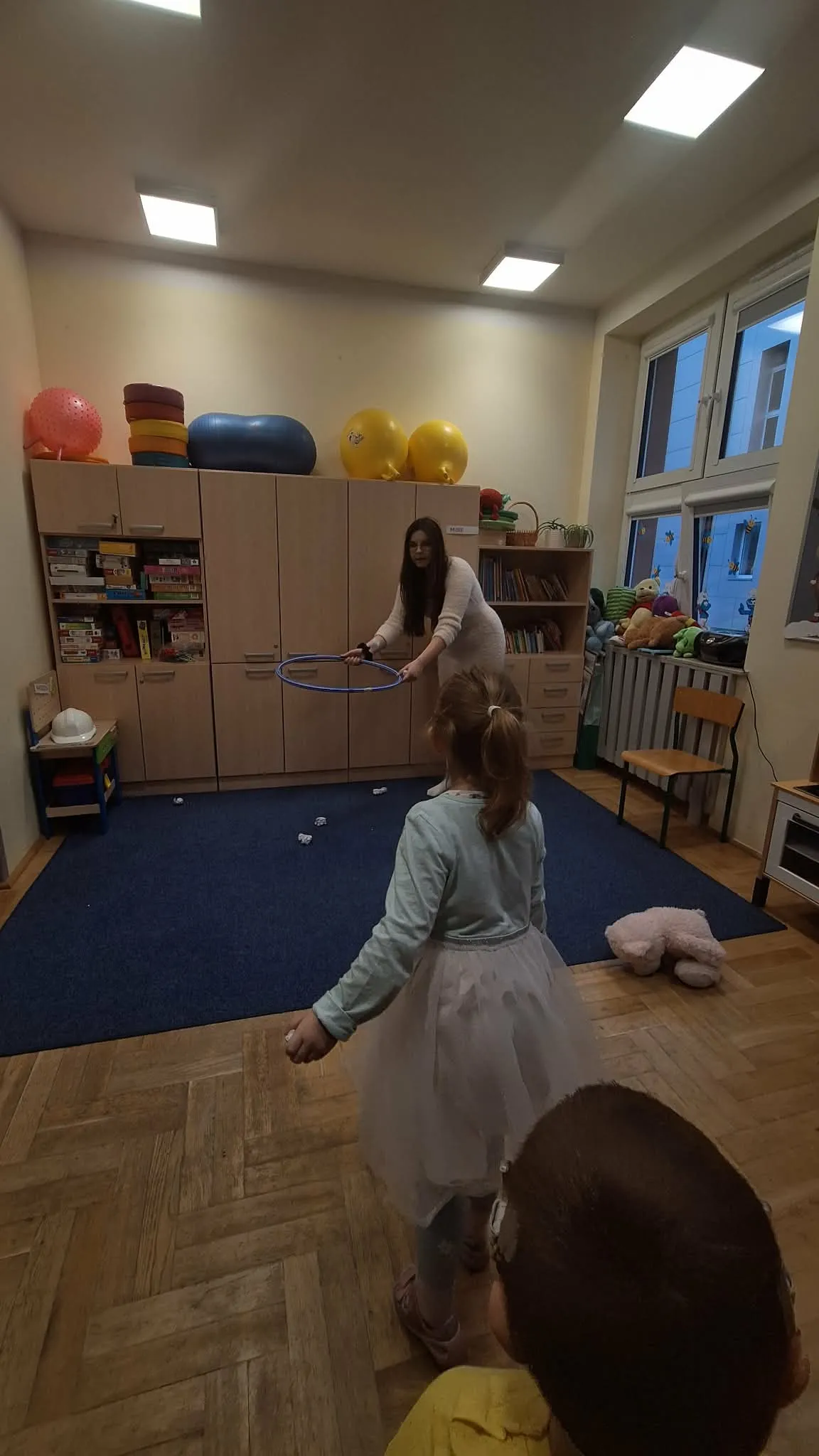 Nauczycielka prowadzi zabawę ruchową z dziećmi w sali, używając obręczy jako elementu ćwiczeń.