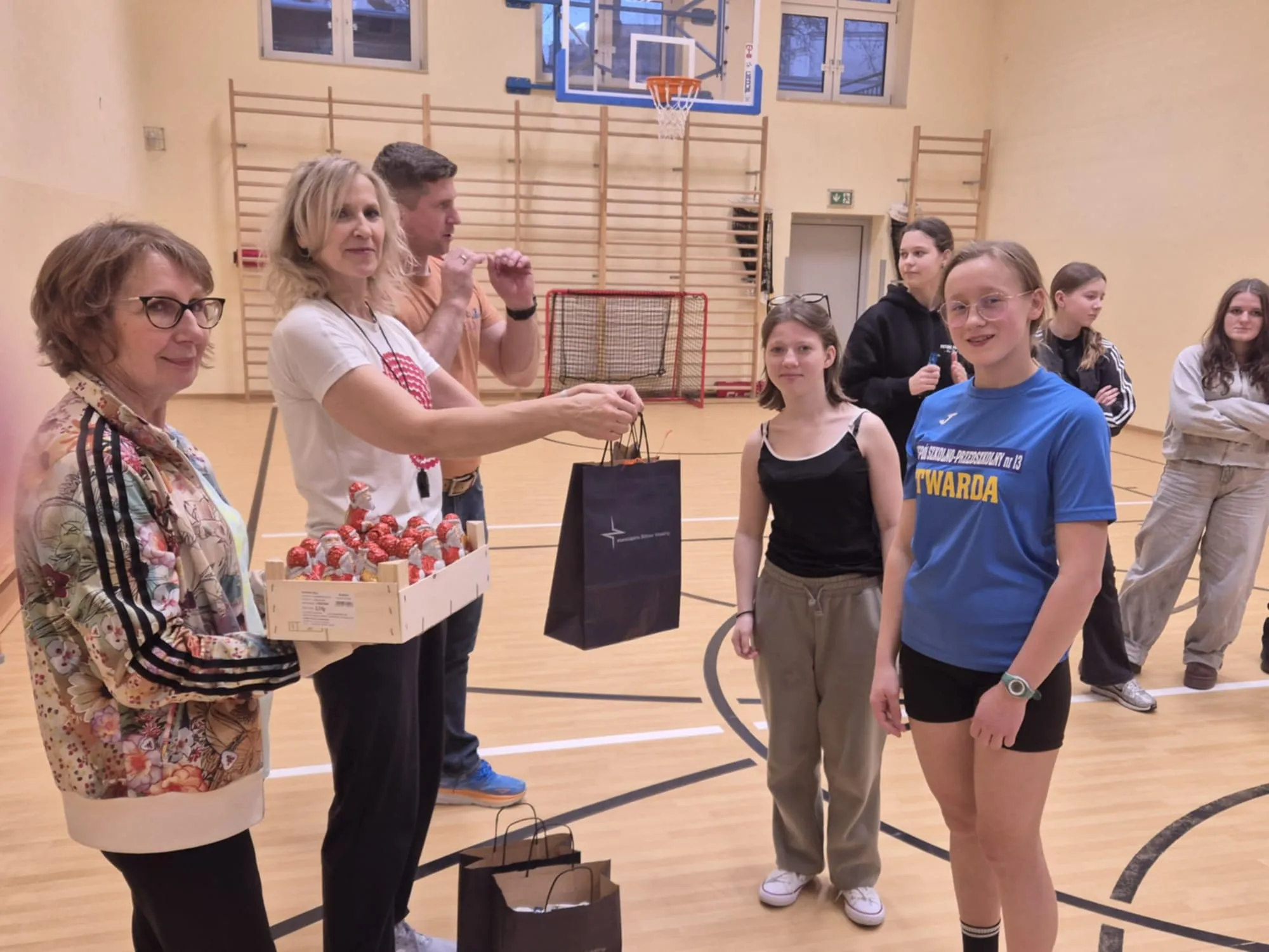 Wręczanie nagród uczennicom w szkolnej sali gimnastycznej, nauczyciele stoją naprzeciw grupy młodzieży, widoczne upominki i kosz z czekoladkami.