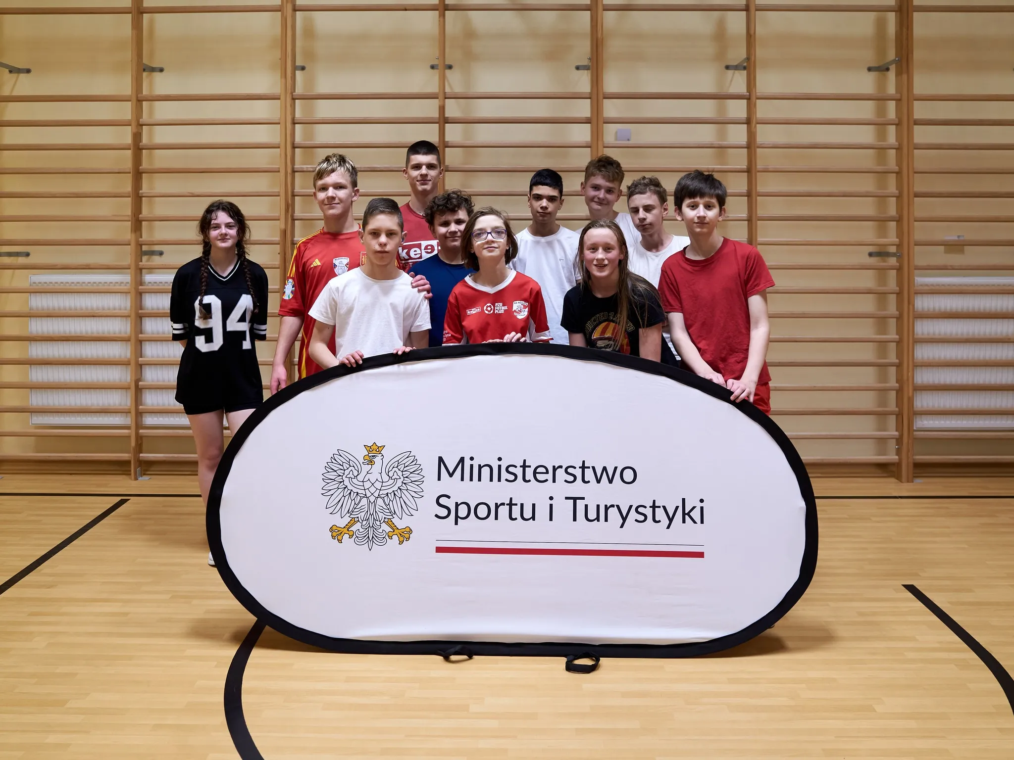 „Grupa młodzieży w sali gimnastycznej stojąca za banerem Ministerstwa Sportu i Turystyki; uczestnicy w strojach sportowych i codziennych, pozują do wspólnego zdjęcia.”