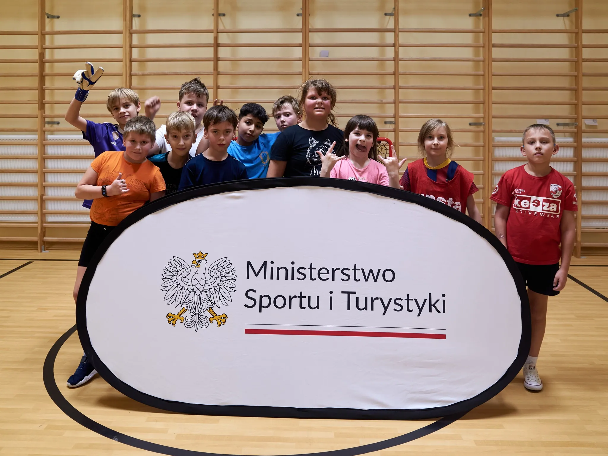 „Grupa dzieci w różnym wieku w sali gimnastycznej, ustawiona za banerem Ministerstwa Sportu i Turystyki; dzieci w kolorowych koszulkach sportowych, pozujące do zdjęcia.”