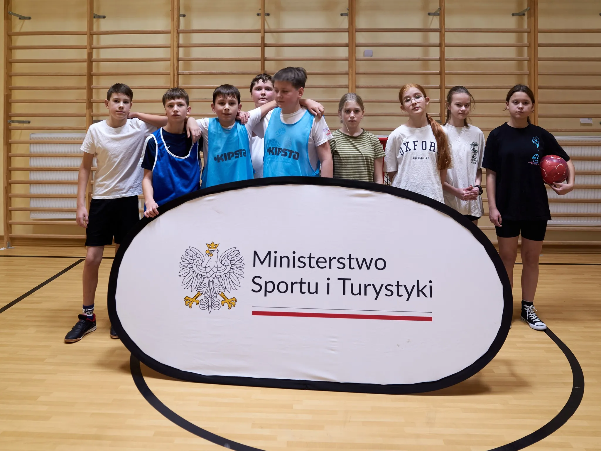„Grupa dzieci w wieku szkolnym stojąca w sali gimnastycznej, pozująca do zdjęcia za banerem Ministerstwa Sportu i Turystyki; część dzieci w strojach sportowych, jedna osoba trzyma piłkę.”