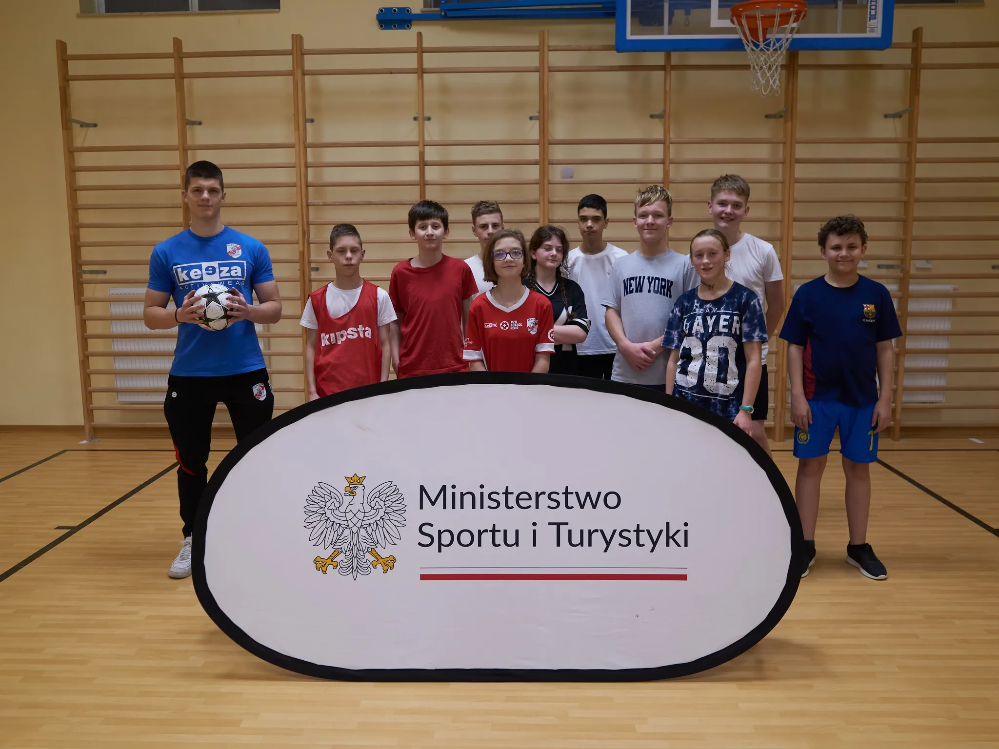 „Instruktor oraz grupa starszych dzieci i młodzieży w sali gimnastycznej, ustawieni za banerem Ministerstwa Sportu i Turystyki; uczestnicy ubrani w koszulki sportowe i codzienne.”
