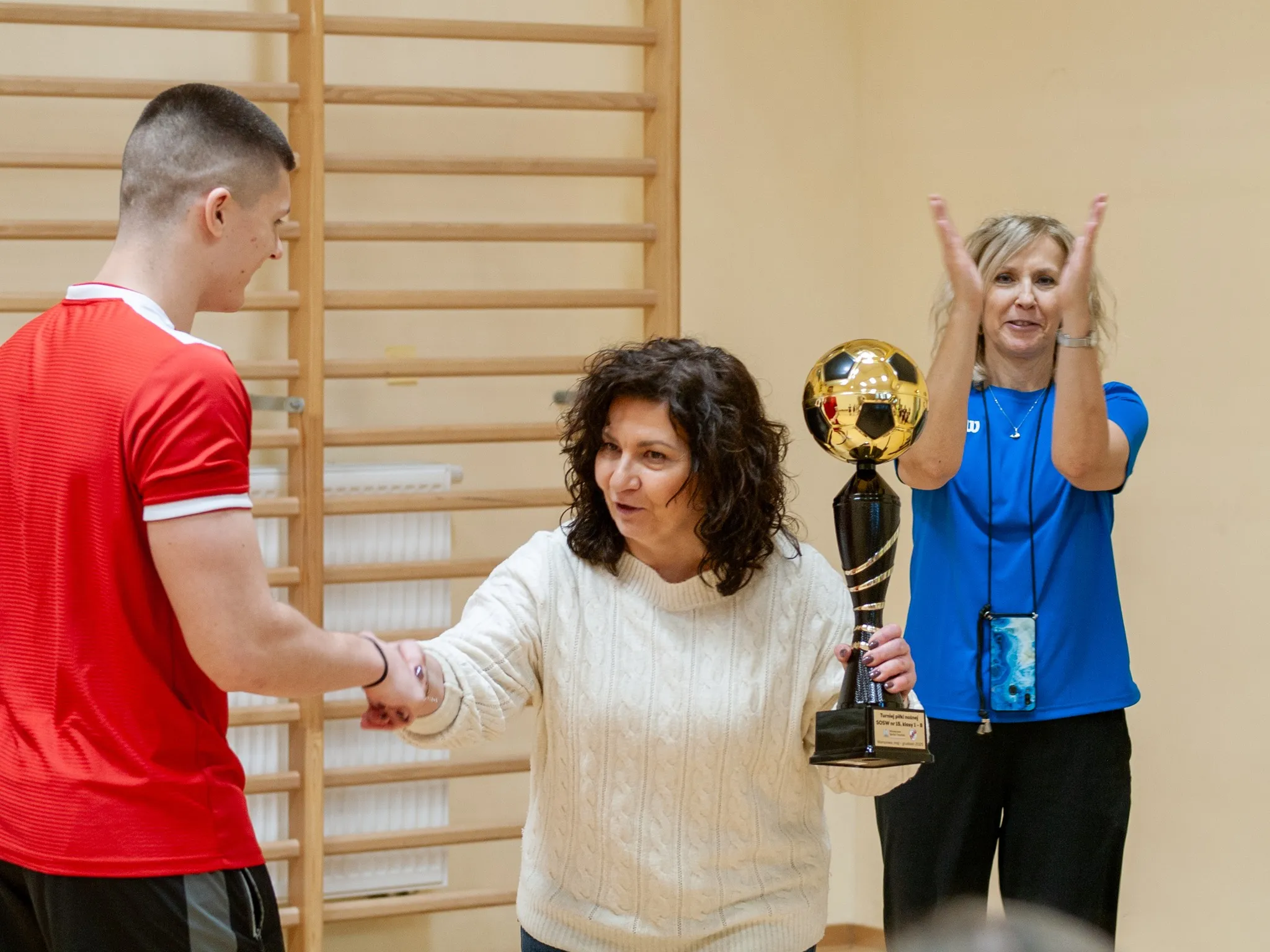 „Wręczenie pucharu – kobieta odbiera puchar, podając trenerowi w czerwonej koszulce rękę; obok osoba bijąca brawo.”