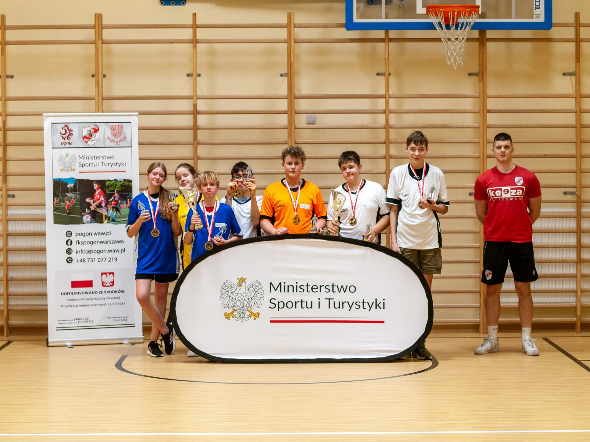 „Grupa młodzieży z medalami i pucharami w sali gimnastycznej, stojąca za banerem Ministerstwa Sportu i Turystyki; obok trener.”