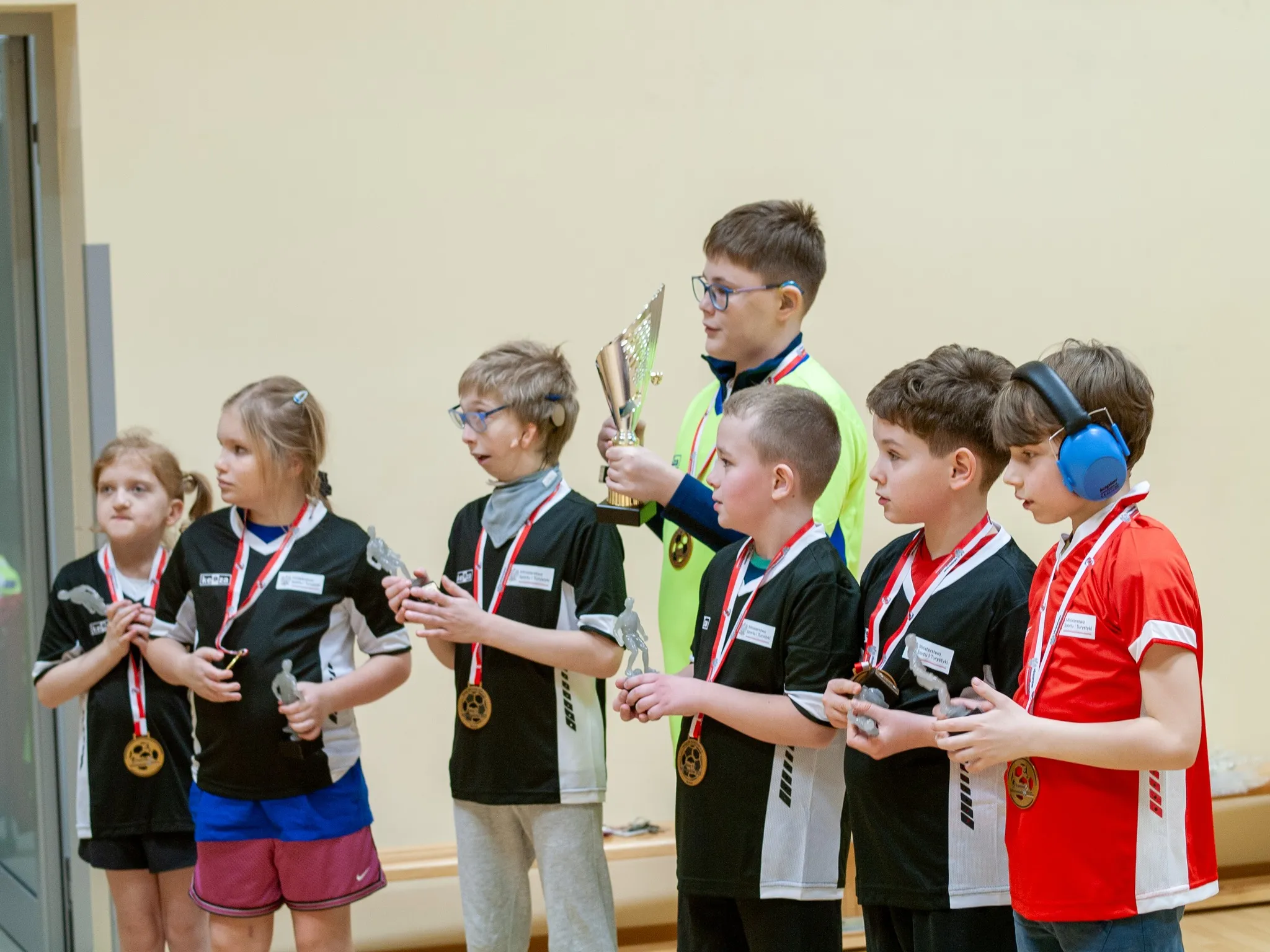 „Dzieci ustawione w rzędzie, trzymające statuetki i puchary; część dzieci w koszulkach sportowych, jedno dziecko z ochronnikami słuchu.”