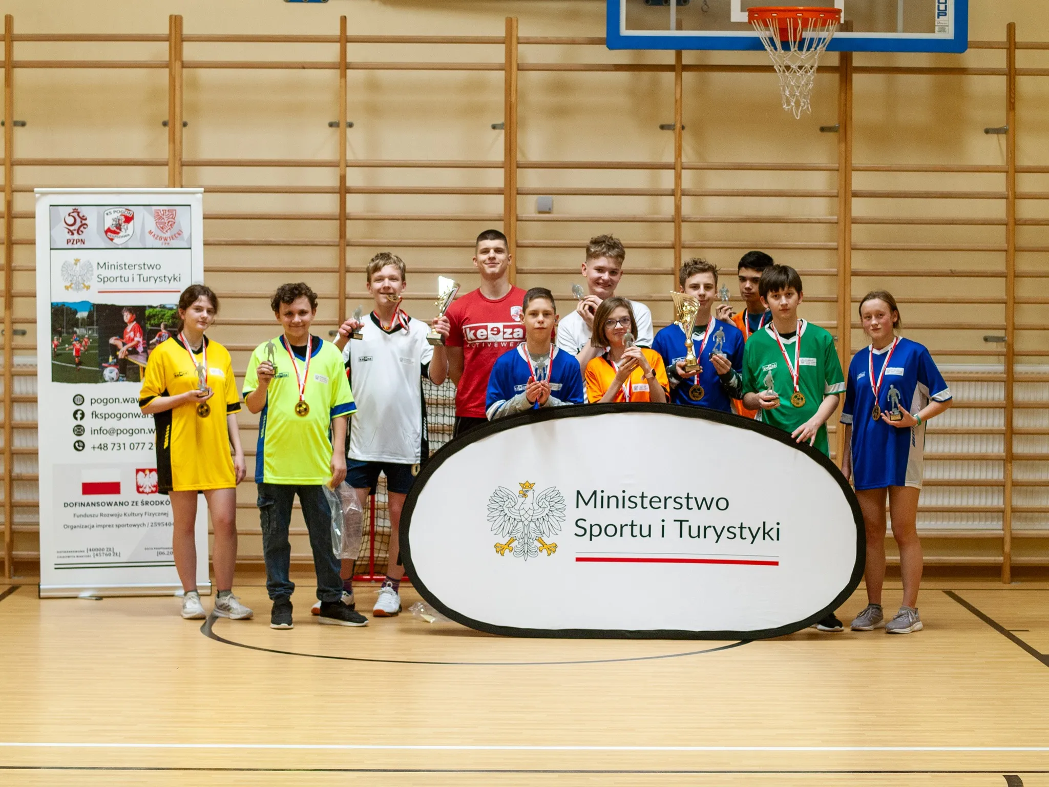 „Grupa młodzieży i trener w sali gimnastycznej, pozujący z medalami i pucharami za banerem Ministerstwa Sportu i Turystyki.”
