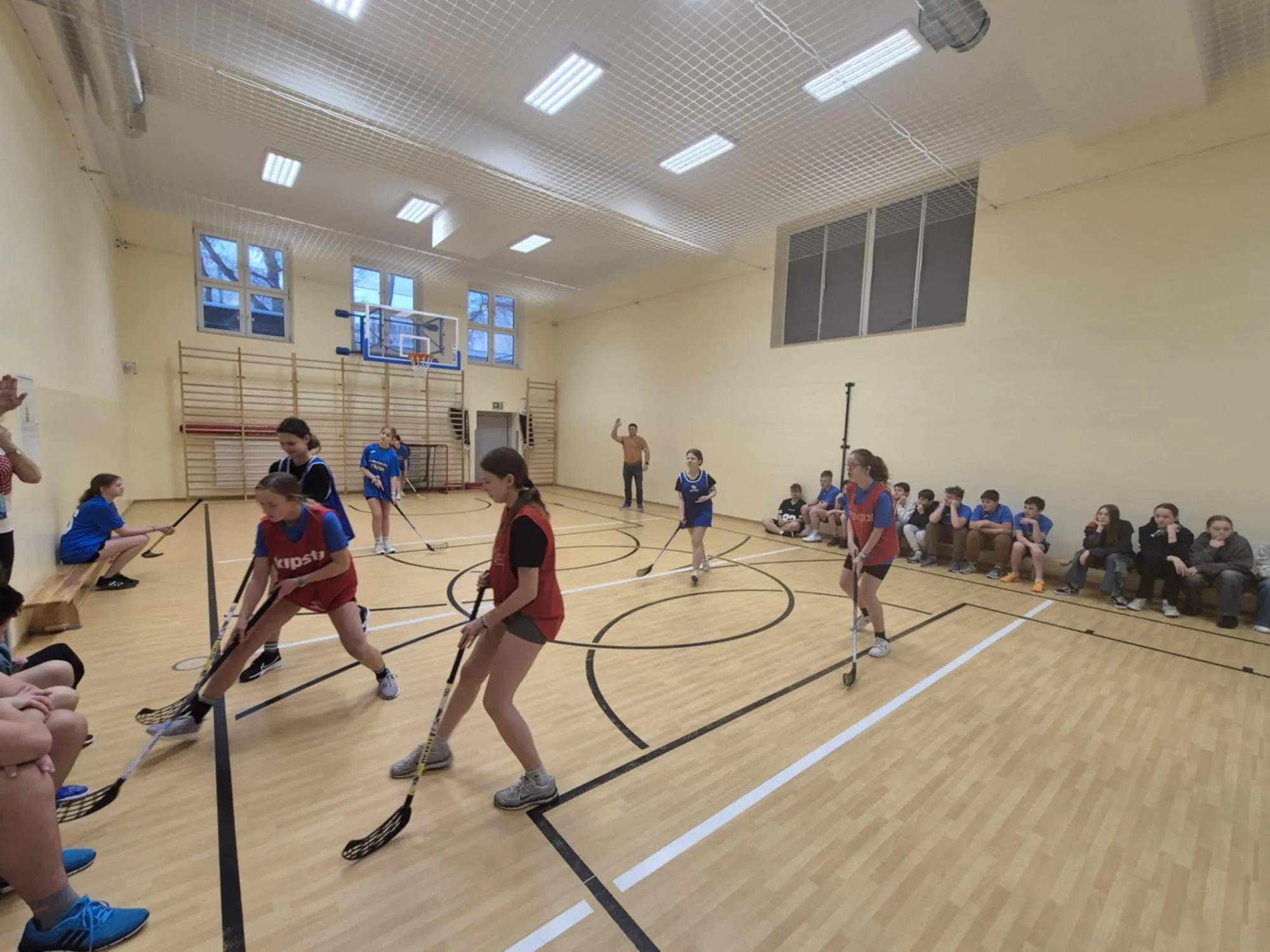 Uczennice w strojach sportowych rywalizują w unihokeju, w tle siedzący na ławkach uczniowie obserwują grę.