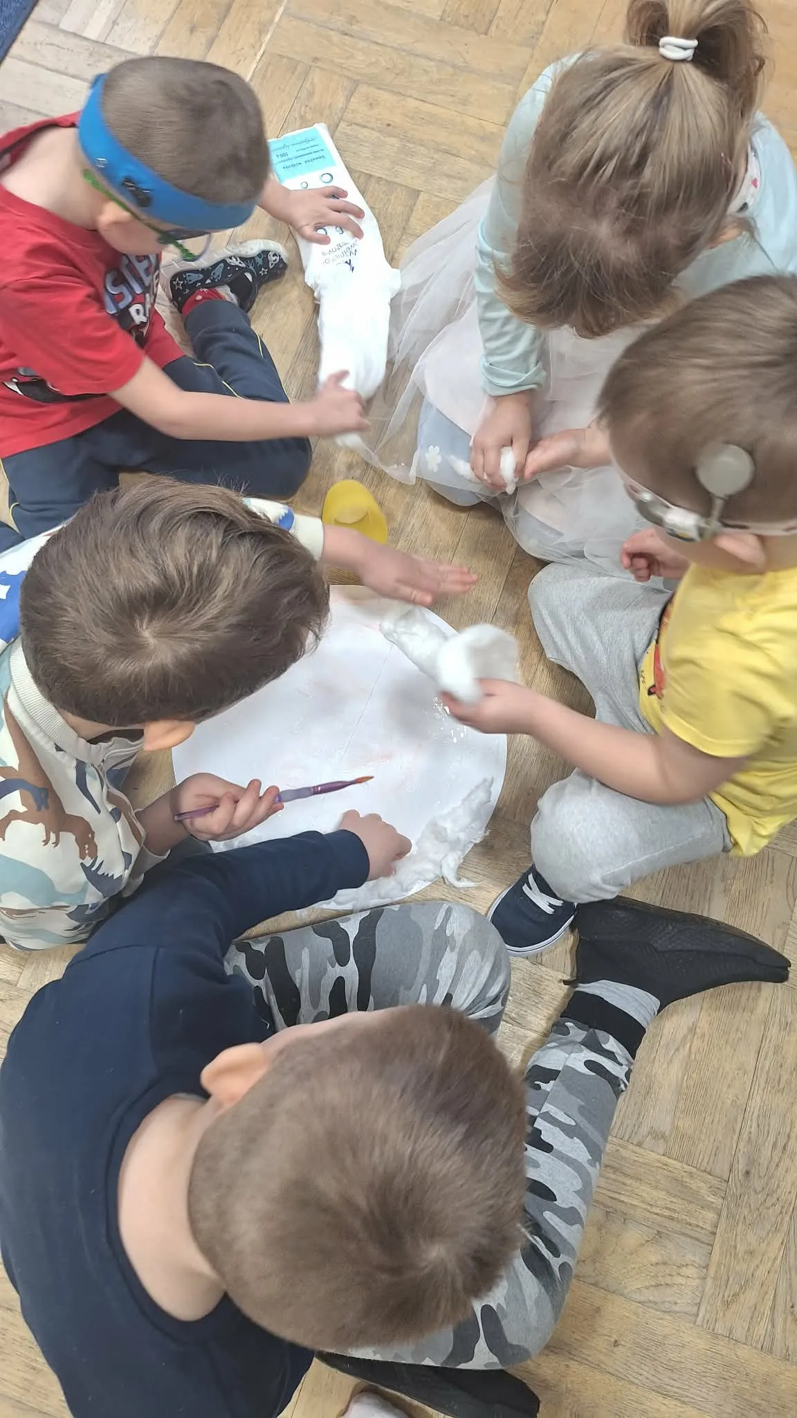 Grupa dzieci siedzi na podłodze i wspólnie wykonuje pracę plastyczną z wykorzystaniem waty oraz farb.