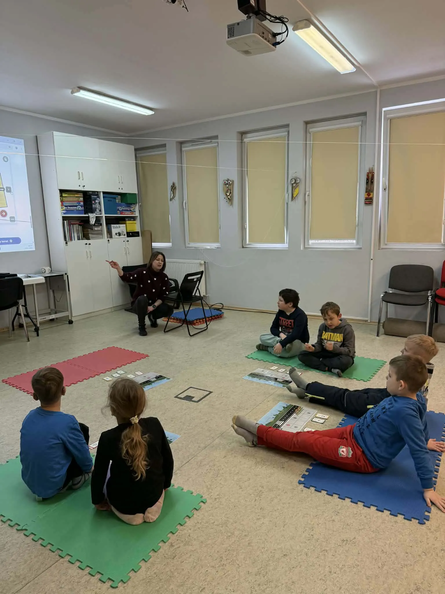 Nauczycielka prowadzi zajęcia z dziećmi siedzącymi na kolorowych matach w sali lekcyjnej. Na podłodze rozłożone są materiały edukacyjne.