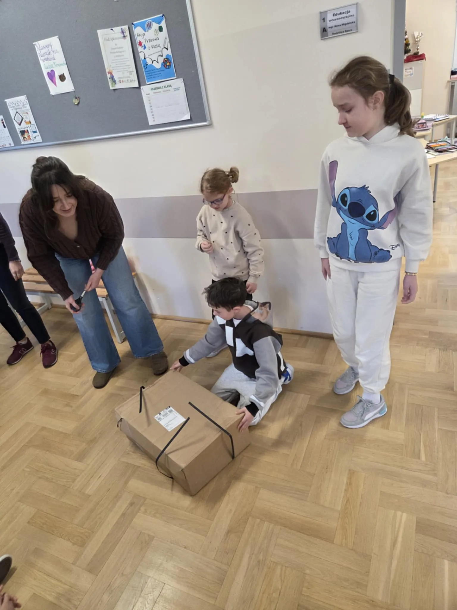 Dzieci i nauczycielka stoją w szkolnym korytarzu, a jedno z dzieci klęczy i rozpakowuje duże kartonowe pudełko leżące na podłodze.