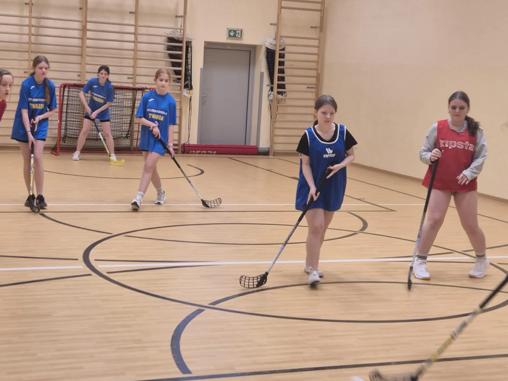 Dziewczęta ćwiczą grę w unihokeja w sali gimnastycznej, każda trzyma kij i ustawia się do zagrania.
