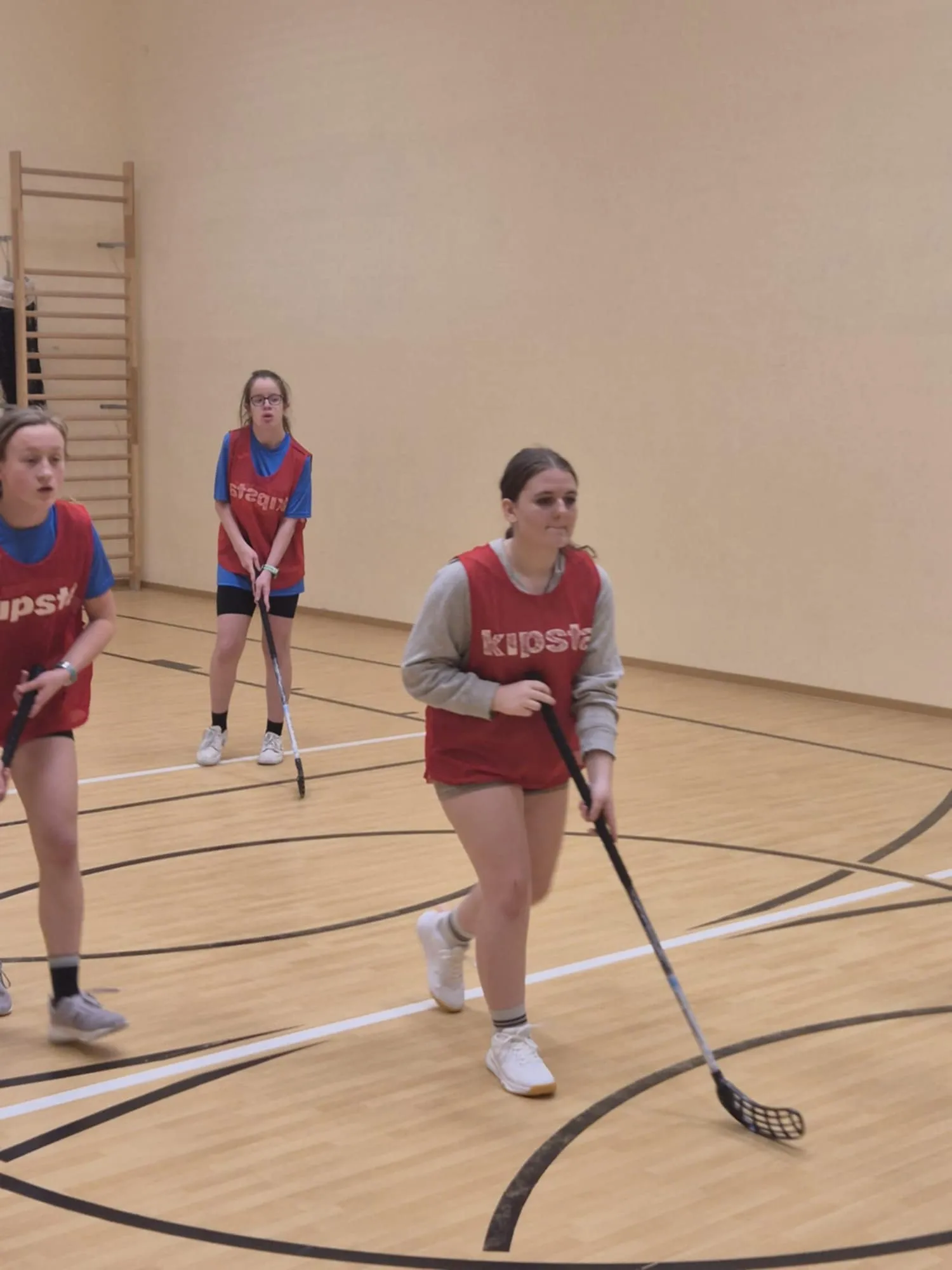 Fragment gry w unihokeja, zawodniczki biegną za piłką, w tle stoją uczniowie kibicujący przy ścianie.