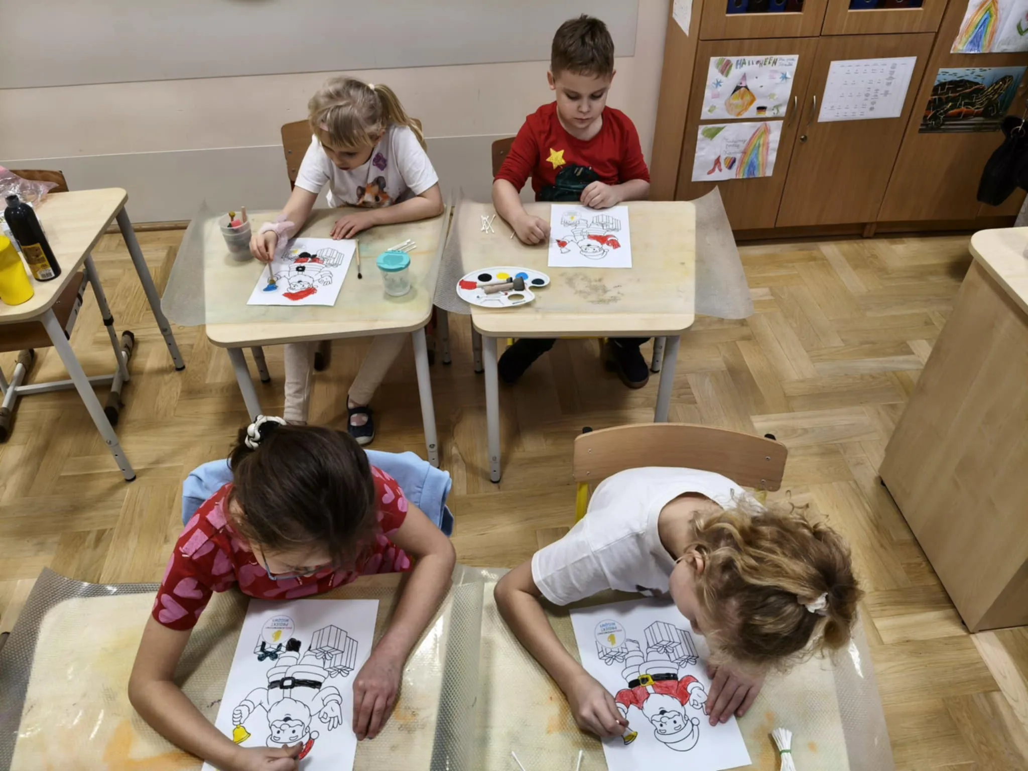 Dzieci siedzą przy stolikach i kolorują przygotowane karty pracy z motywem zimowym, używając farb i kredek.