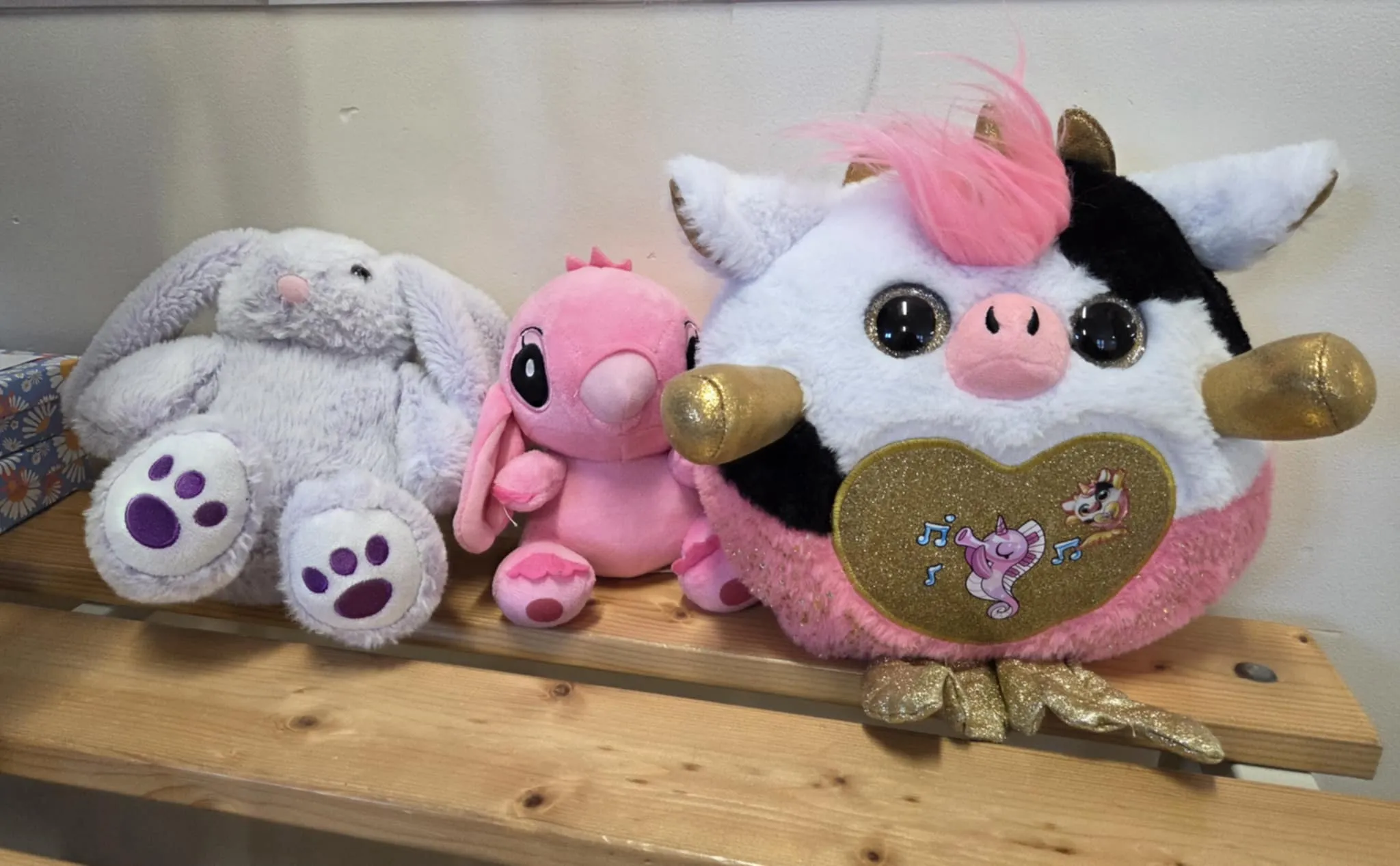 Trzy pluszowe zabawki ustawione na drewnianej ławce: szary królik, różowy króliczek oraz duży pluszowy jednorożec trzymający brokatowe serce.