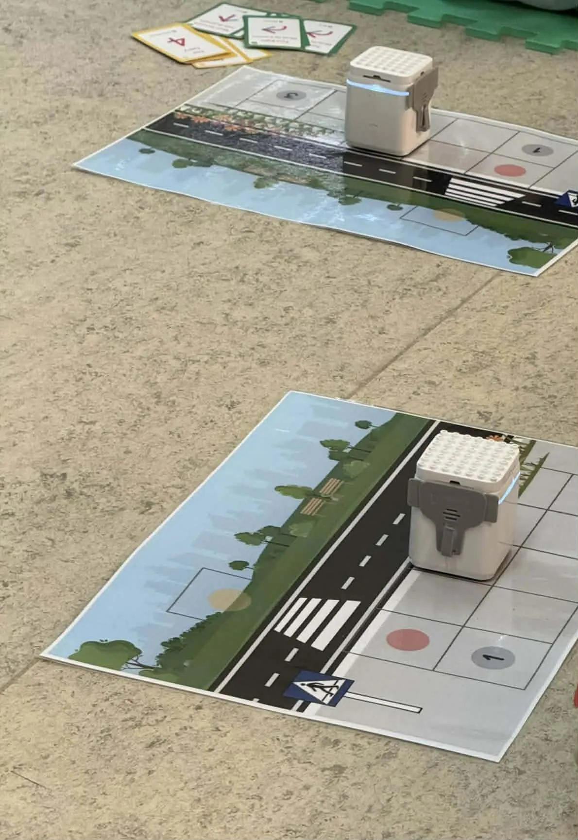 Maty edukacyjne z ilustracją drogi i przejścia dla pieszych, na których ustawione są małe roboty do nauki podstaw programowania i bezpieczeństwa na drodze.