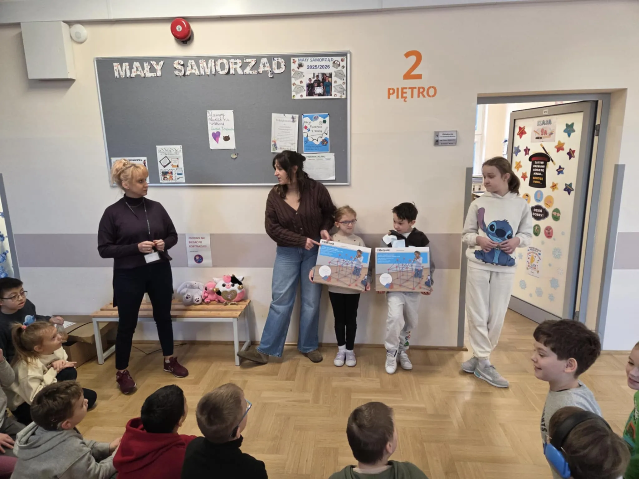 Uczniowie i nauczyciele stoją w szkolnym korytarzu, prezentując dwa pudełka z zestawami konstrukcyjnymi przed grupą siedzących dzieci.