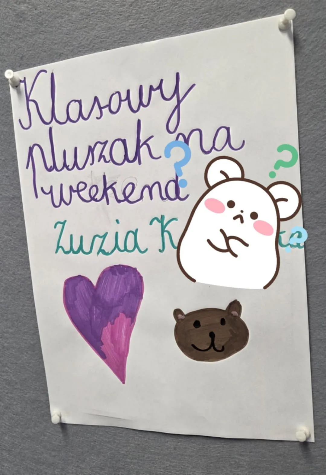 Ręcznie wykonany plakat przypięty do tablicy korkowej z napisem „Klasowy pluszak na weekend”. Na plakacie narysowane są serce oraz głowy misiów w pastelowych kolorach.