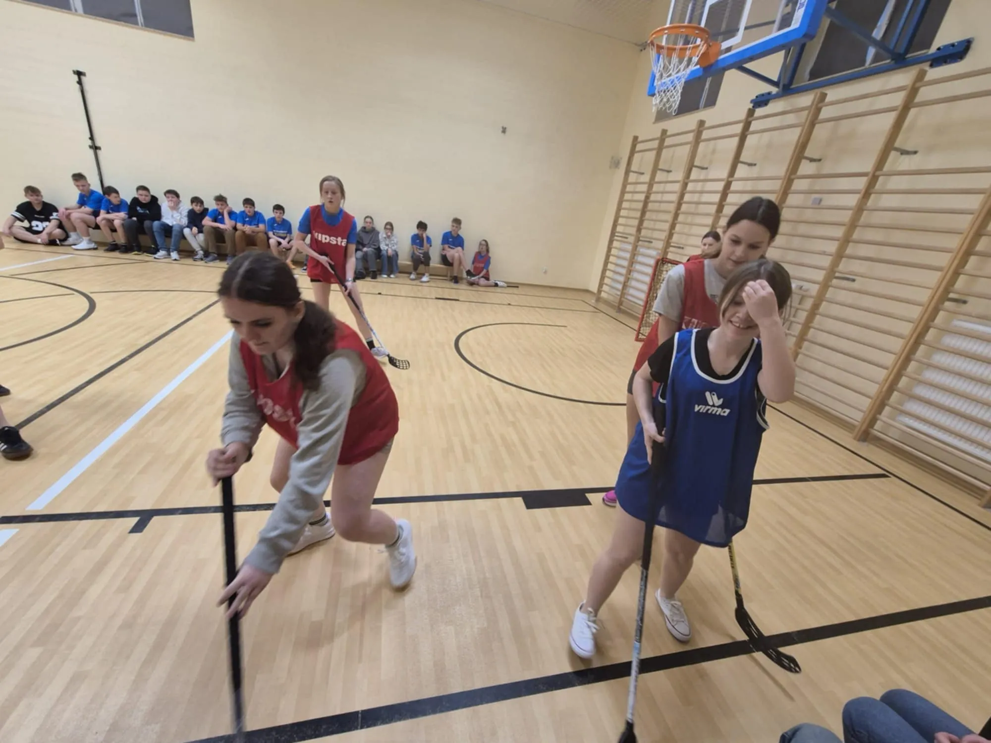 Uczennice w strojach sportowych rywalizują w unihokeju, w tle siedzący na ławkach uczniowie obserwują grę.