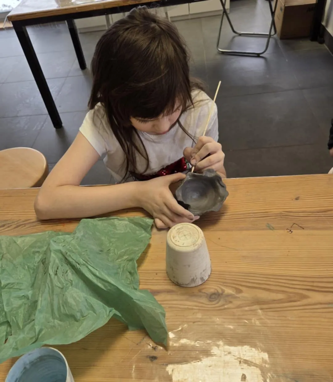 Dziewczynka przy stole precyzyjnie zdobi małą miseczkę z gliny przy pomocy drewnianego patyczka.