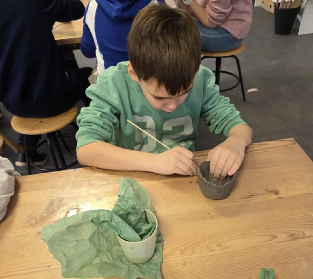 Chłopiec siedzący przy stole wygładza i zdobi małe naczynie z gliny przy użyciu patyczka.