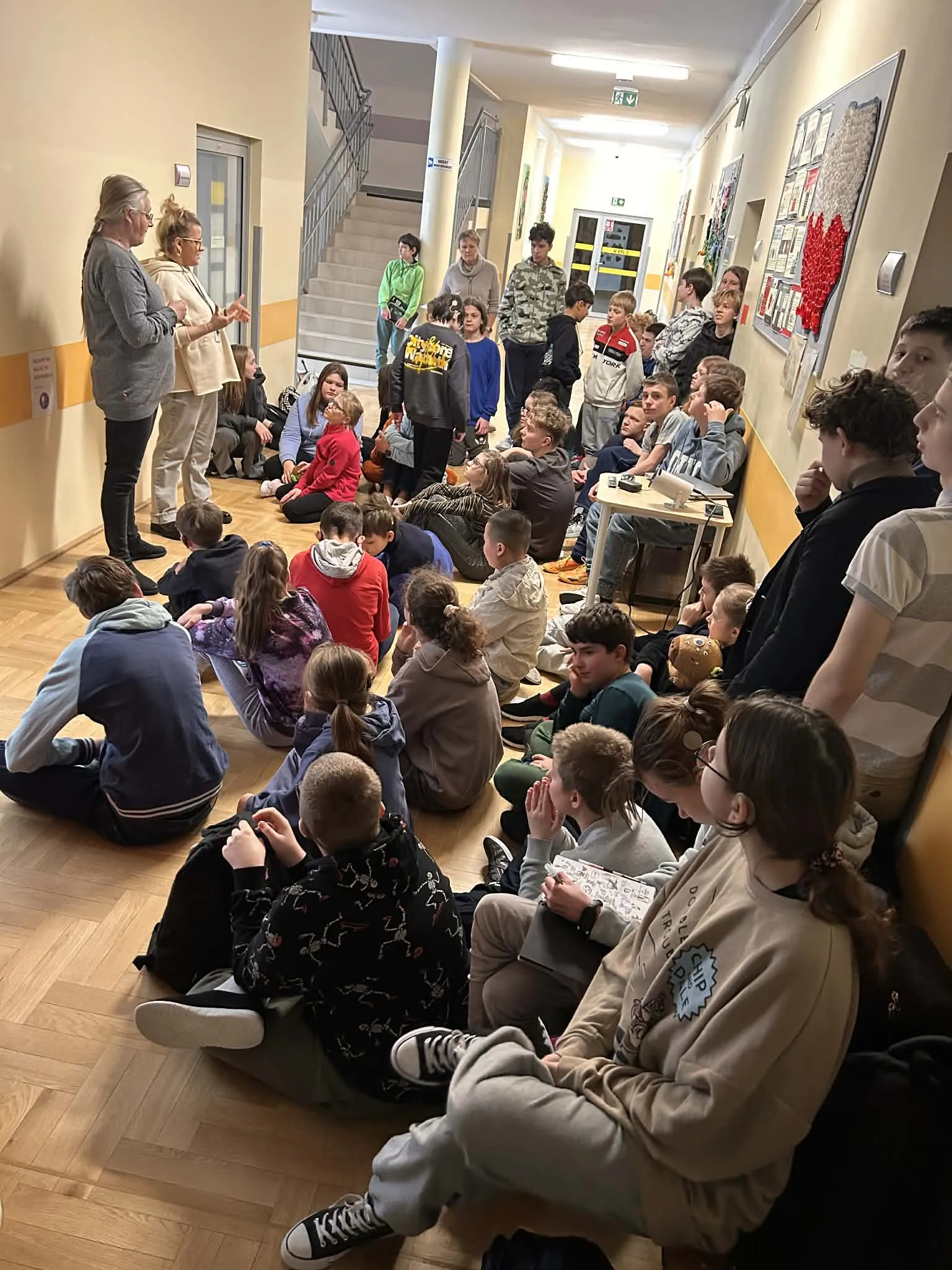 Uczniowie siedzący na korytarzu słuchają pań prowadzących spotkanie