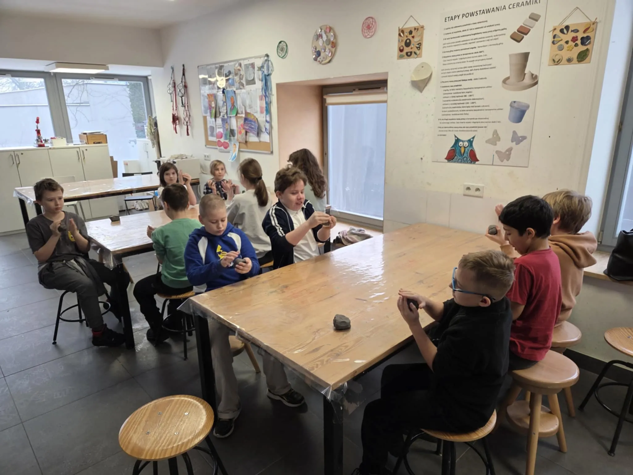 Grupa uczniów siedzi przy stołach w pracowni plastycznej i formuje naczynia z gliny podczas zajęć ceramicznych.