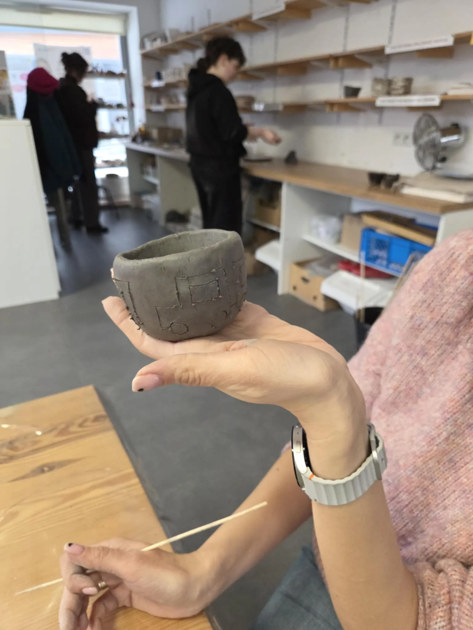 Dłoń trzyma niewielkie naczynie z surowej gliny ozdobione geometrycznym wzorem w pracowni ceramicznej.