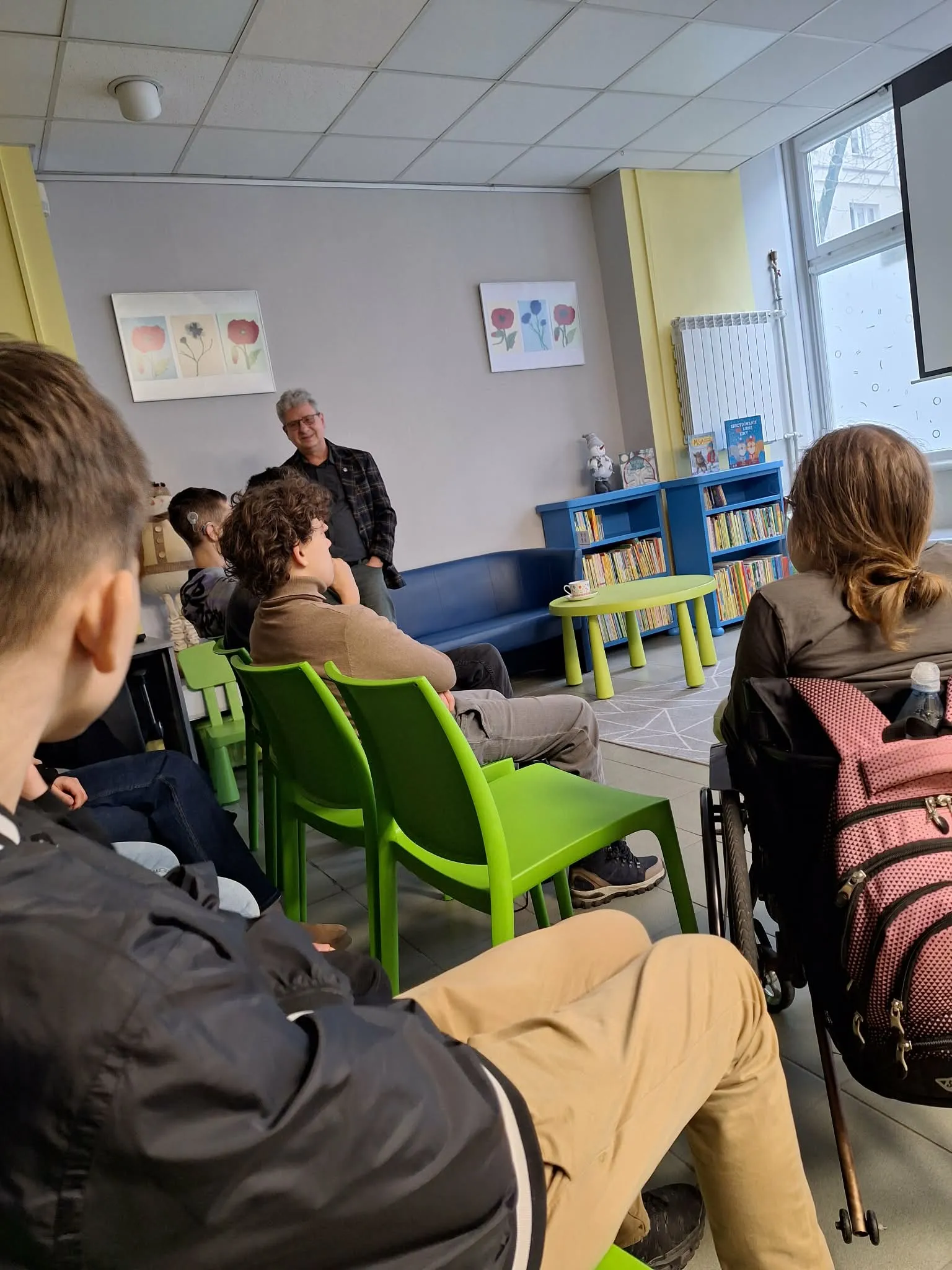 Uczniowie siedzą na zielonych krzesłach i rozmawiają z prowadzącym podczas spotkania edukacyjnego w szkolnej bibliotece.