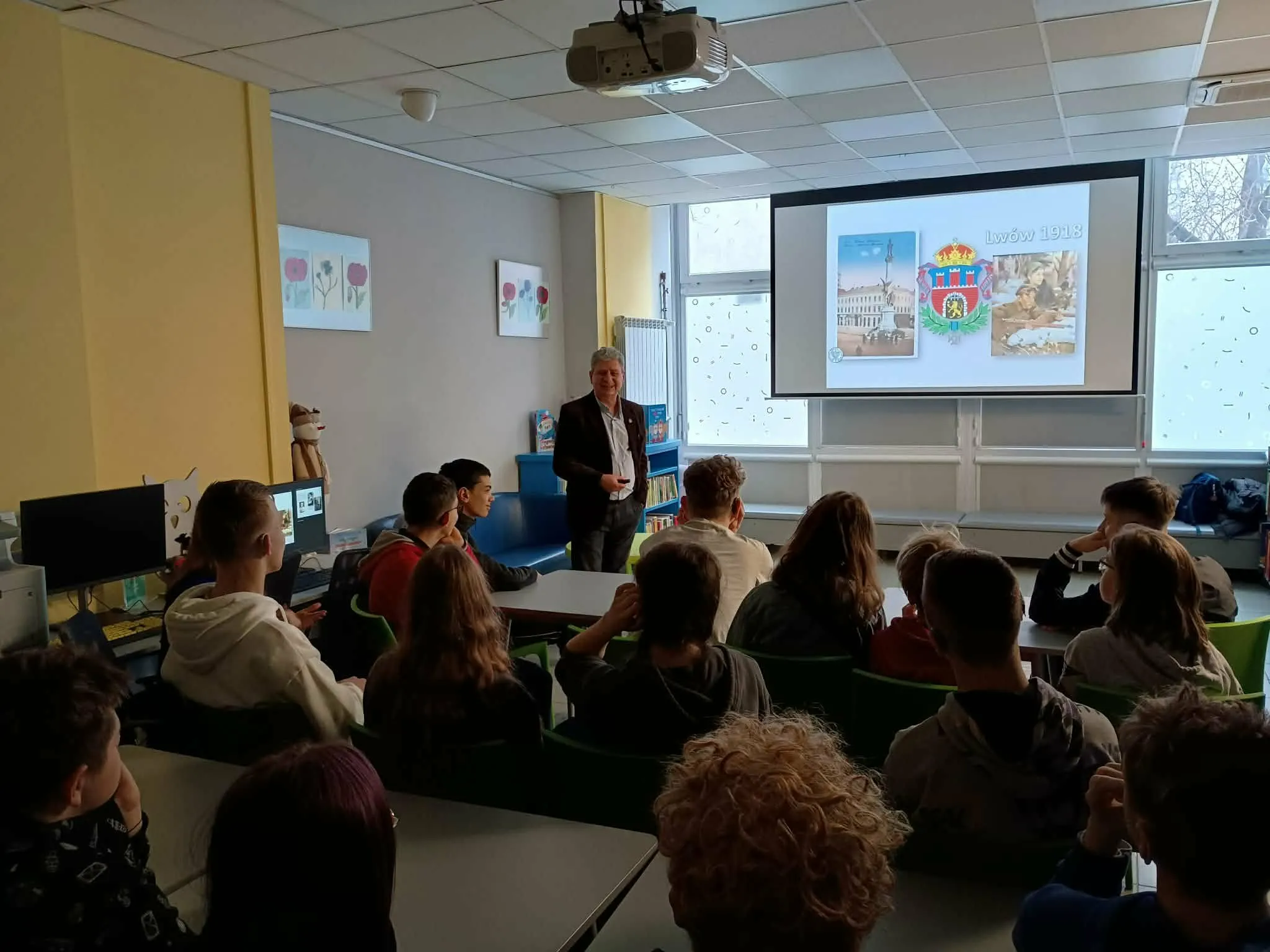 Prelegent prowadzi prezentację historyczną dla uczniów siedzących w sali lekcyjnej przed ekranem z rzutnika.