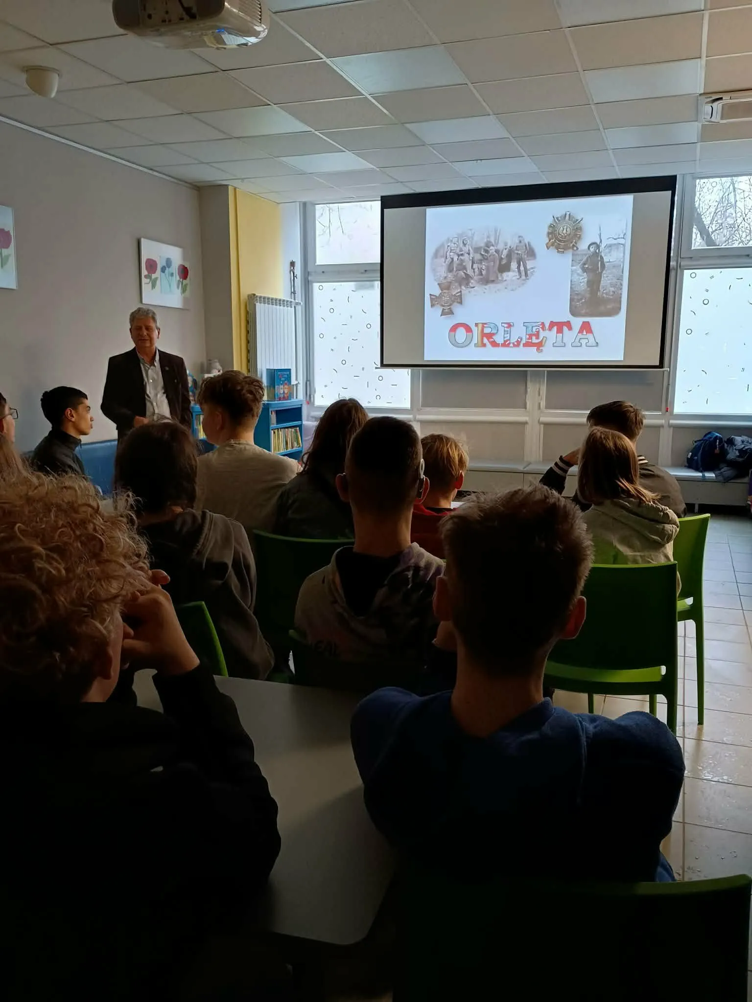 Prelegent prowadzi prezentację historyczną dla uczniów siedzących w sali lekcyjnej przed ekranem z rzutnika.