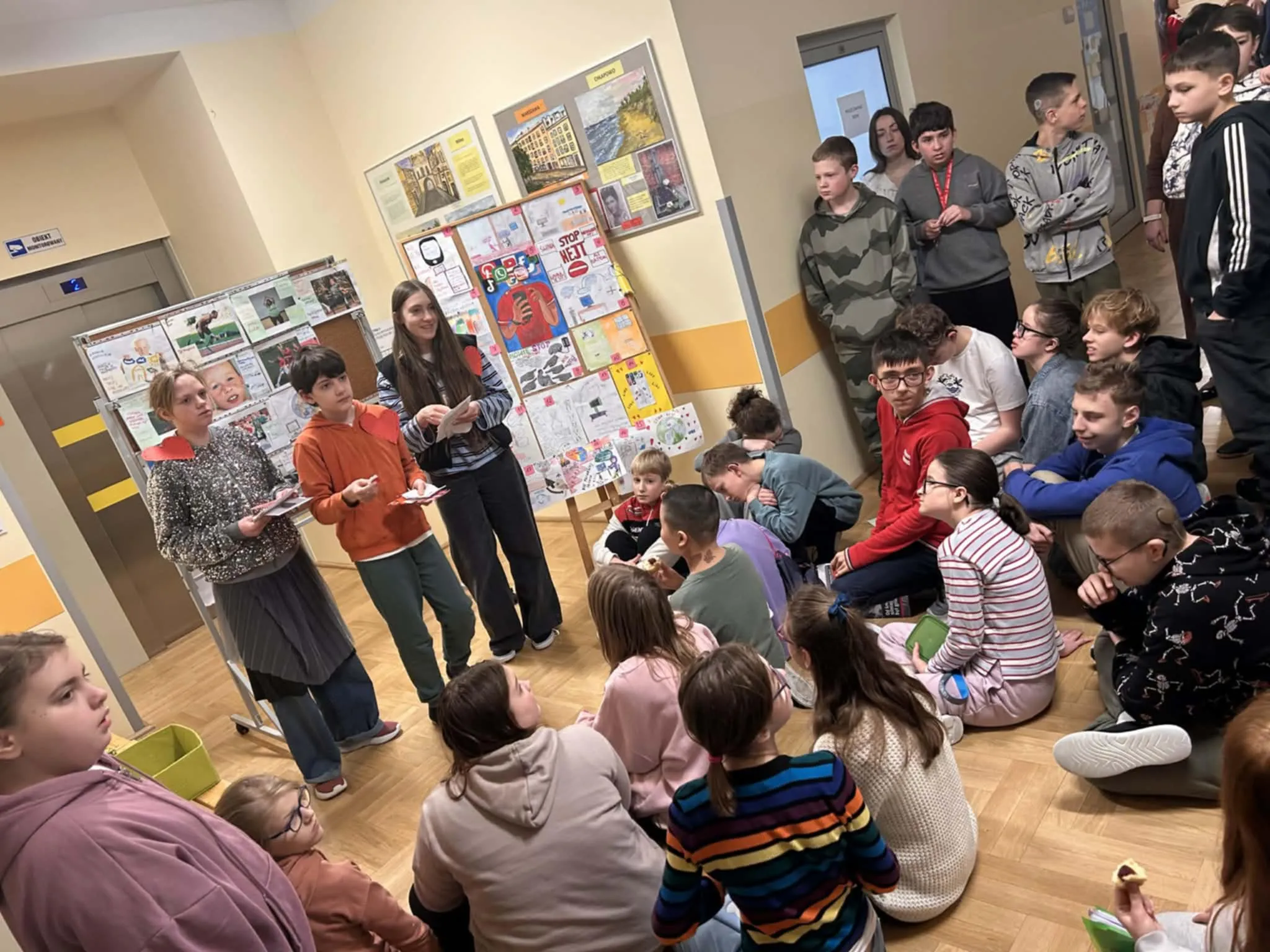 Grupa uczniów zgromadzona na korytarzu szkolnym podczas rozdawania walentynek, w tle tablice z pracami plastycznymi.