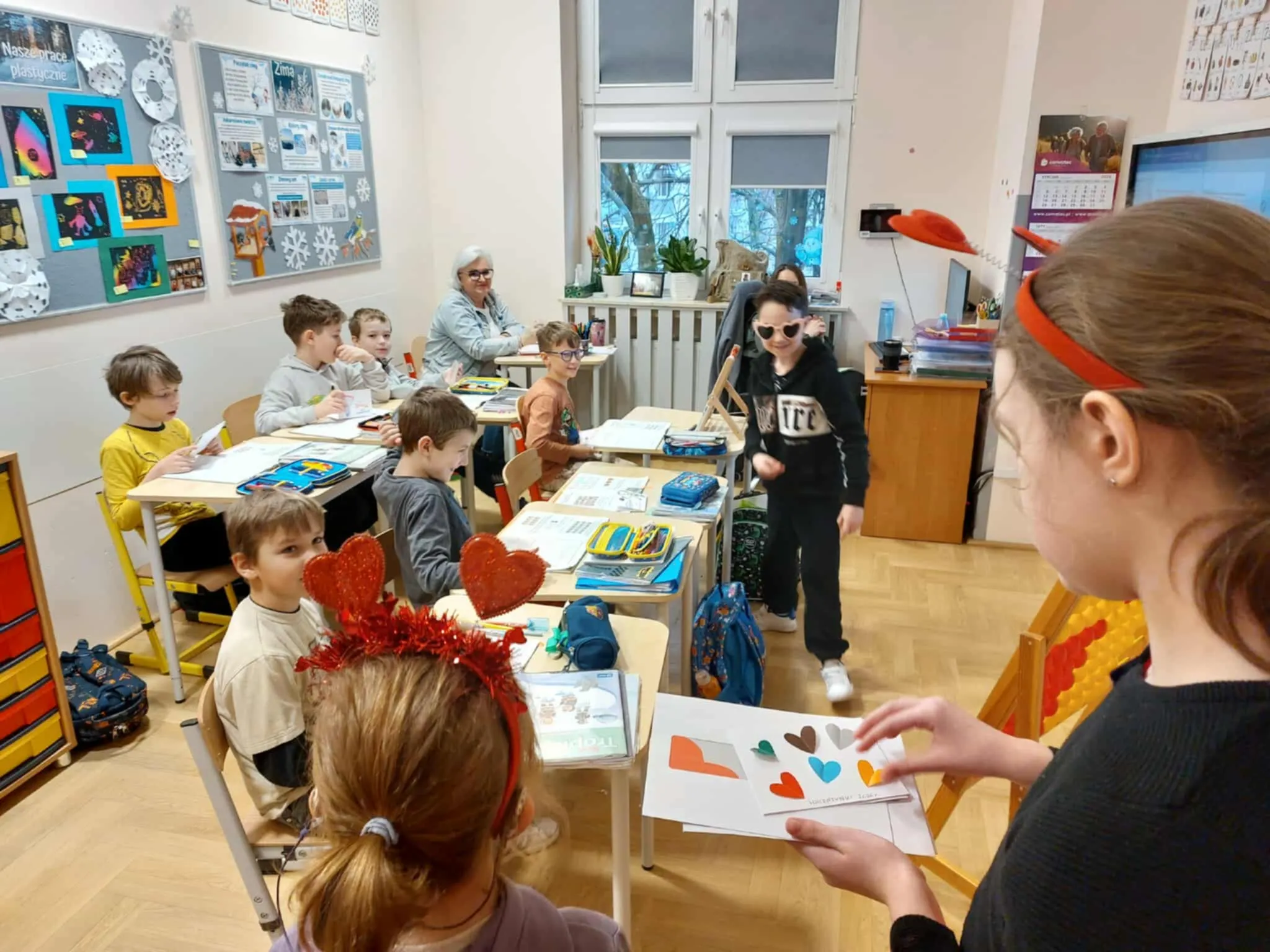Uczniowie siedzą w ławkach podczas lekcji, a walentynkowy listonosz rozdaje kartki w klasie udekorowanej sercami.
