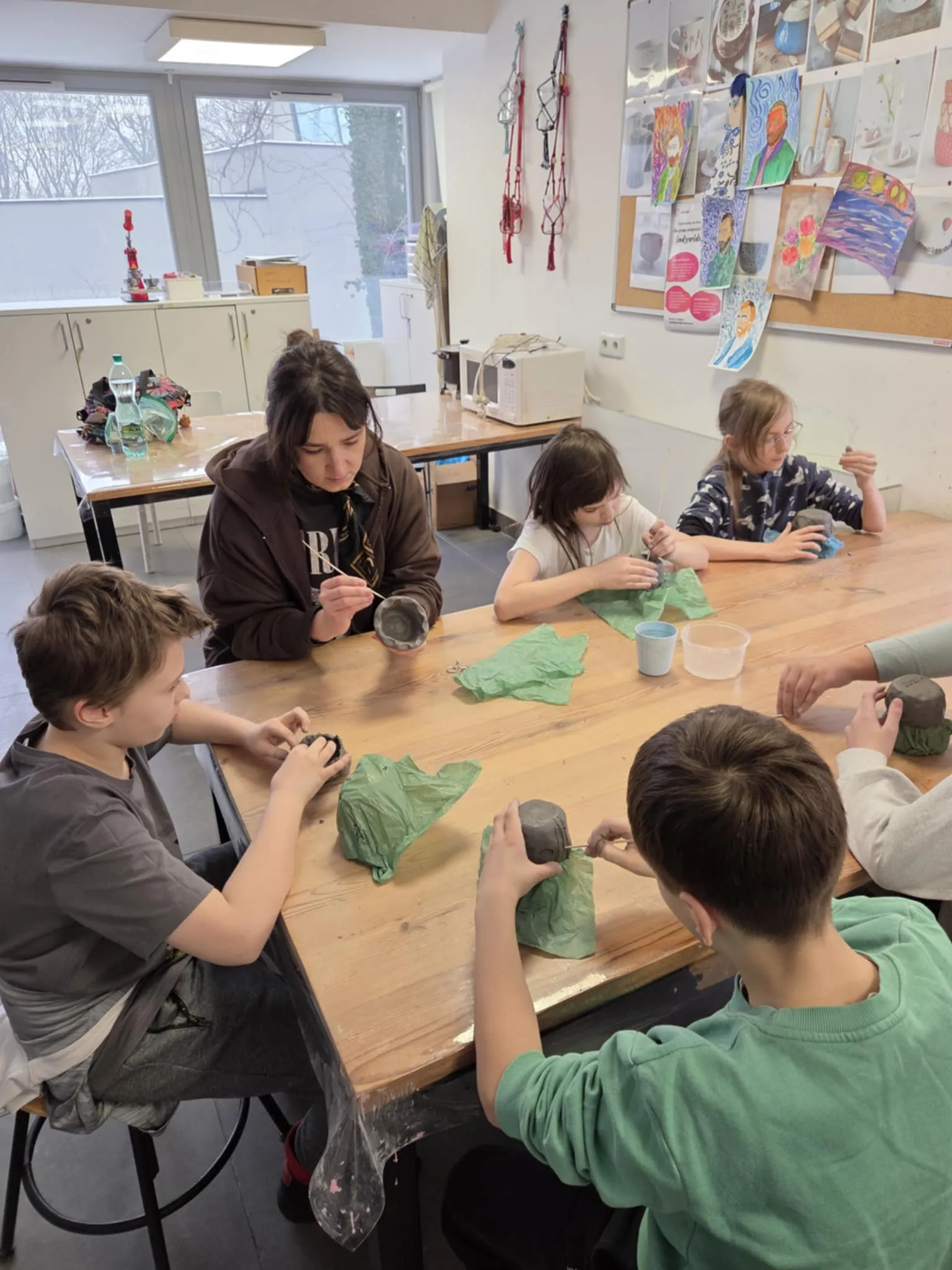 Uczniowie wraz z nauczycielką siedzą przy stole i starannie formują oraz zdobią swoje prace ceramiczne.