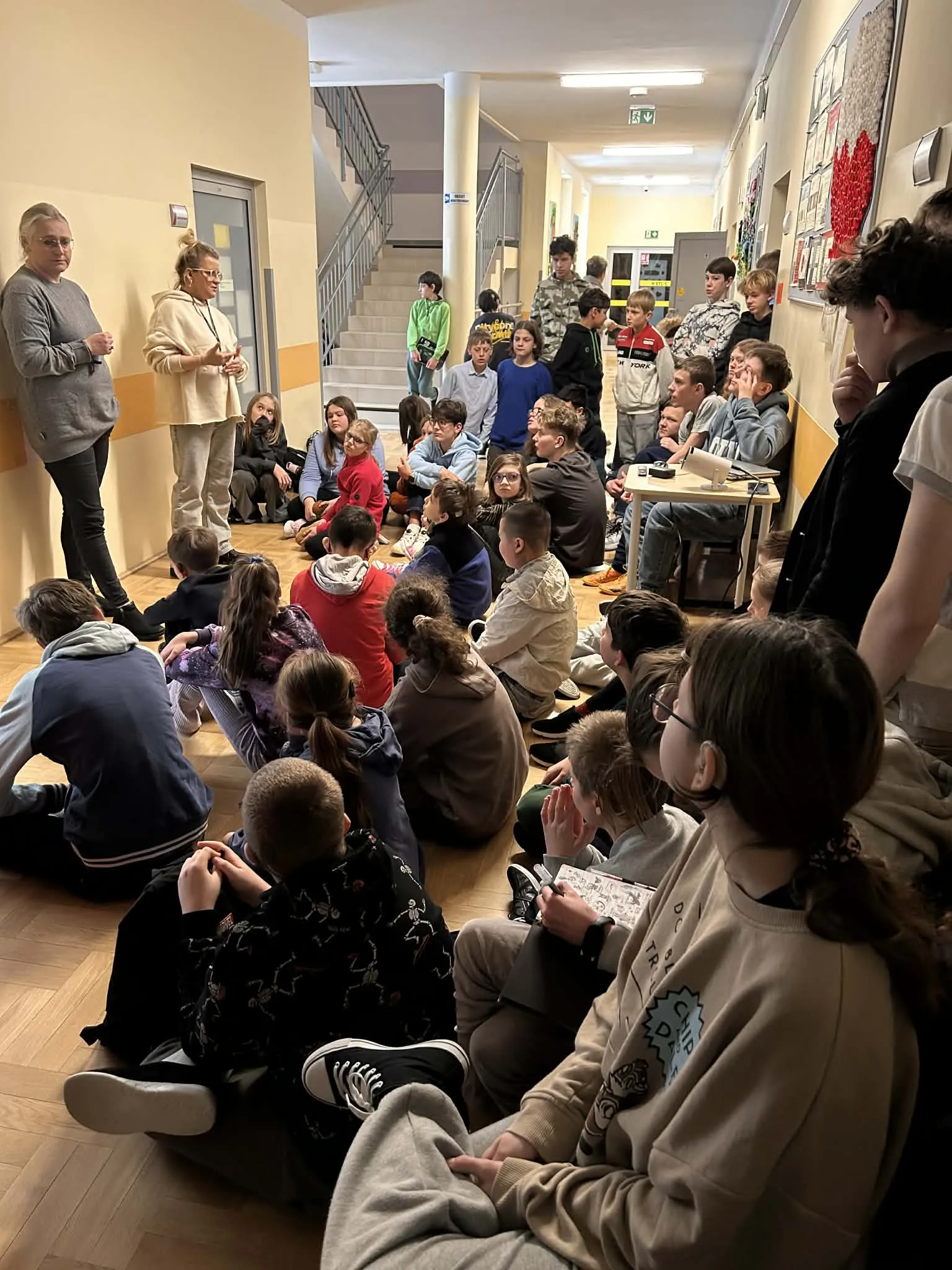 Uczniowie siedzący na korytarzu słuchają pań prowadzących spotkanie