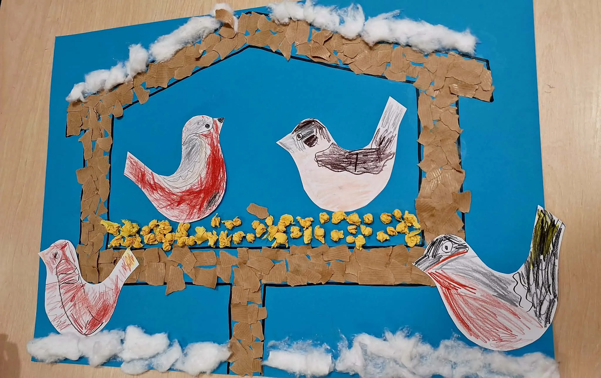 Kolorowa praca plastyczna przedstawiająca ptaki w karmniku wykonana z papieru, waty i bibuły.