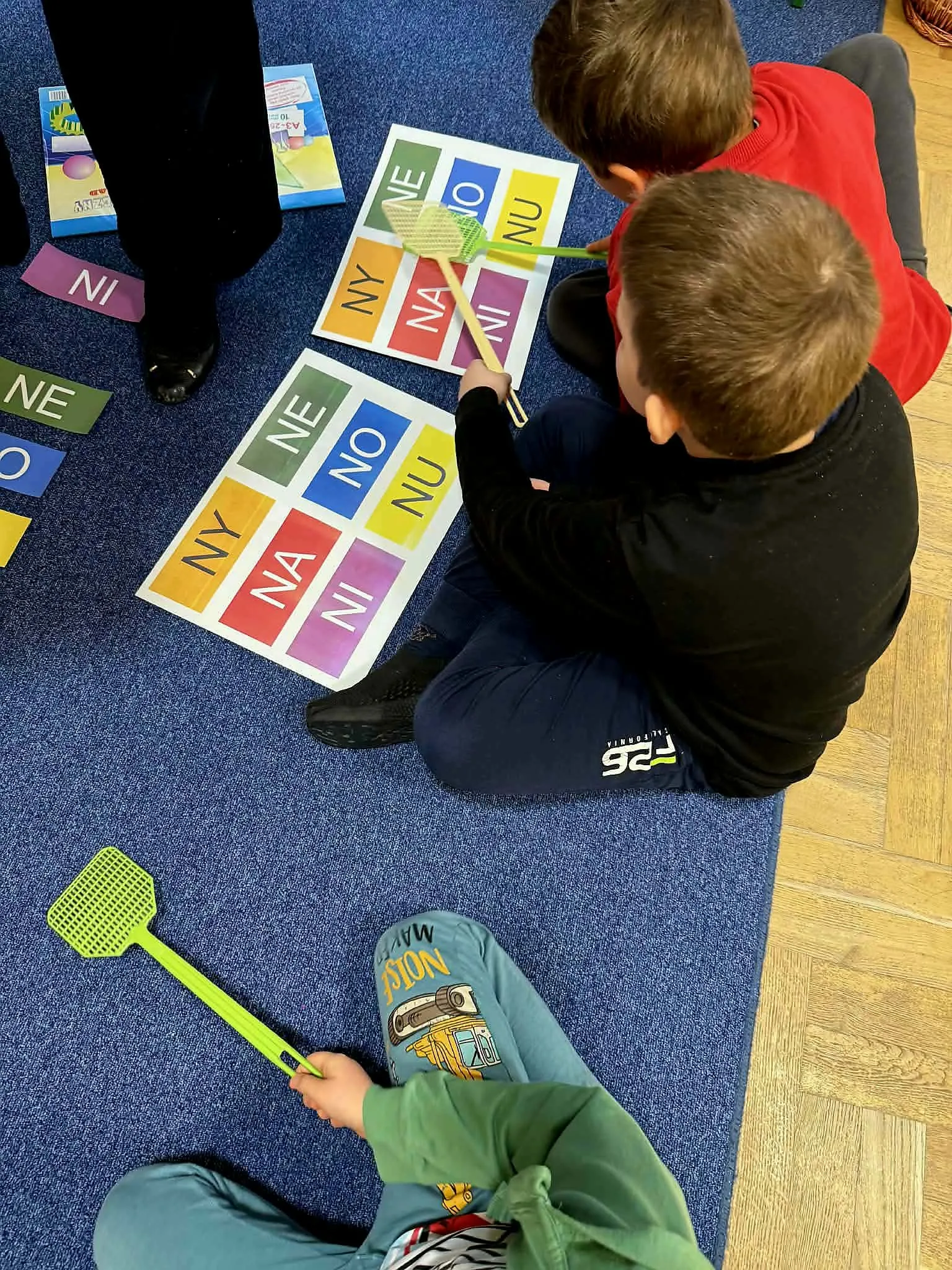 Dwoje dzieci wskazuje packami na plansze z sylabami podczas zajęć edukacyjnych na dywanie.