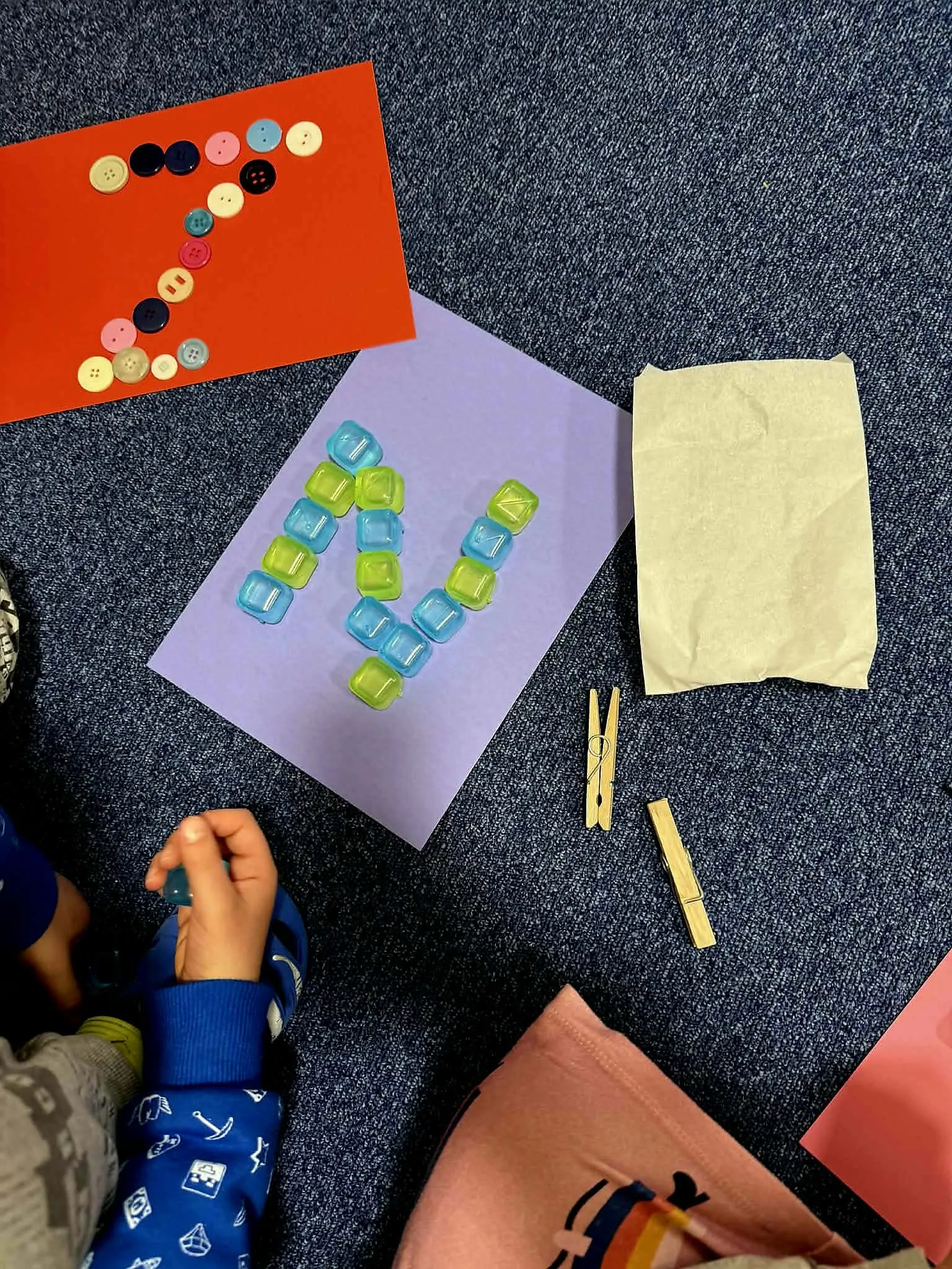 Dzieci tworzą literę N z kolorowych guzików i przezroczystych klocków na kartkach papieru.