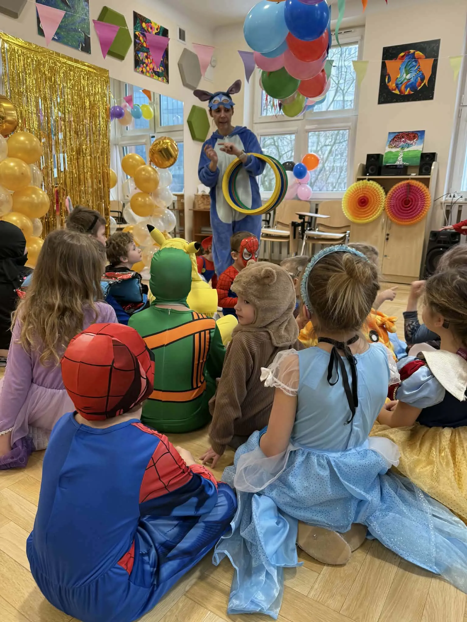 Animator w stroju zwierzątka prowadzi zabawę z obręczami dla dzieci siedzących w przebraniach.