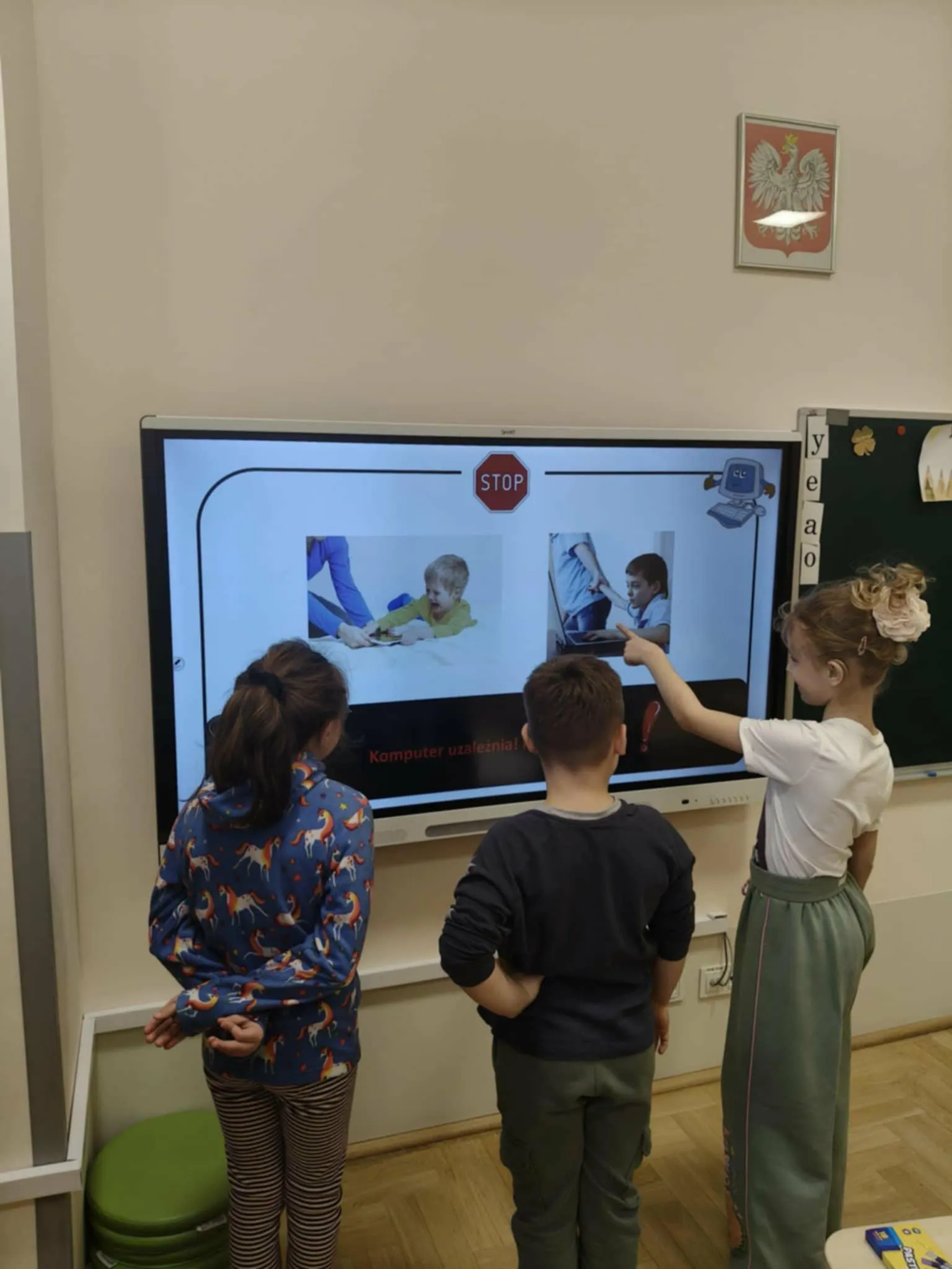 Troje uczniów stoi przy tablicy multimedialnej i wskazuje elementy prezentacji o uzależnieniu od komputera.