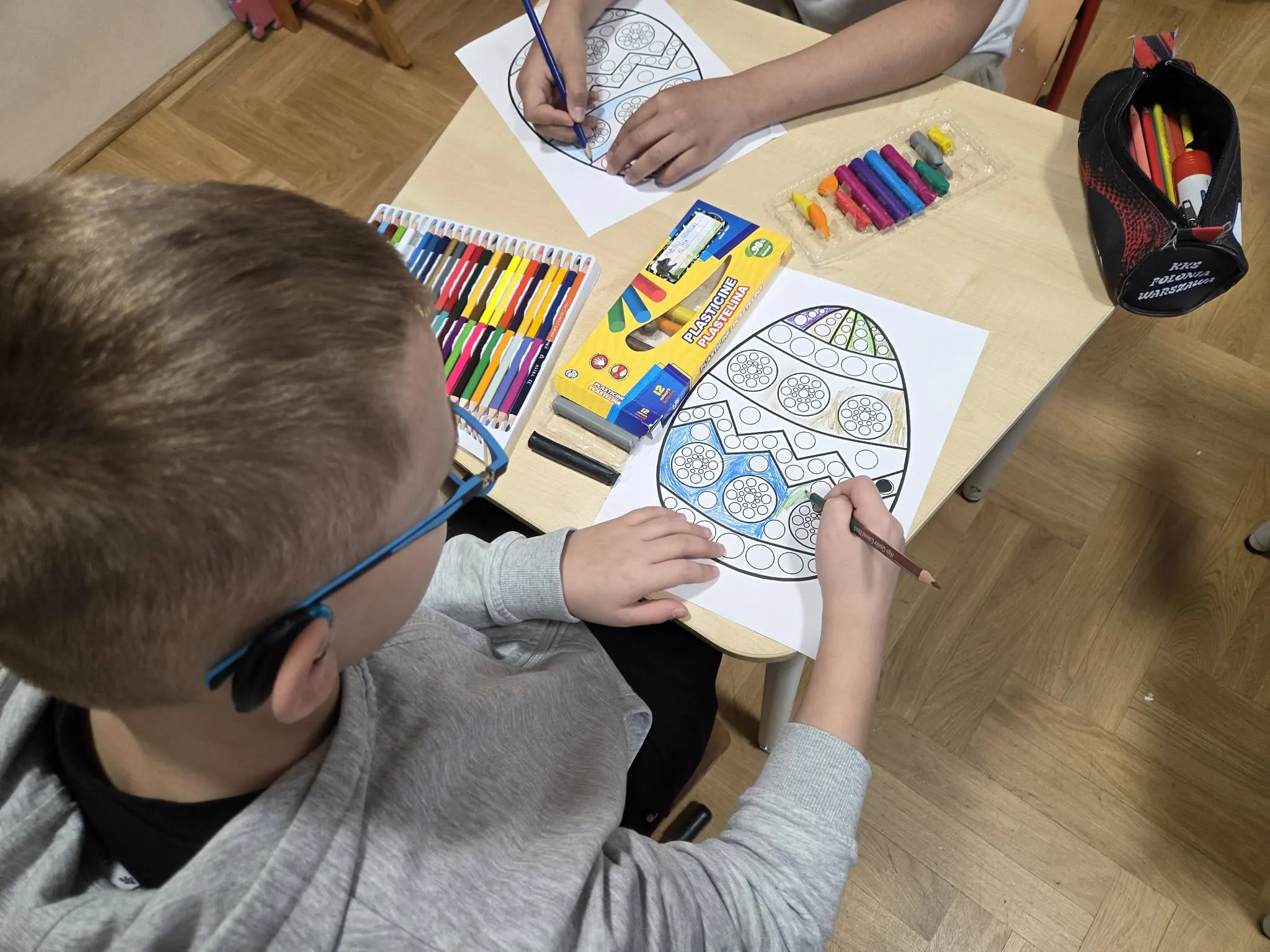 Uczeń koloruje wzór jajka kredkami przy biurku szkolnym