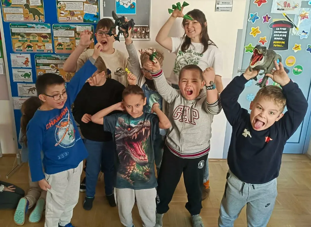 Grupa uczniów wraz z nauczycielką pozują z figurkami dinozaurów w sali lekcyjnej.