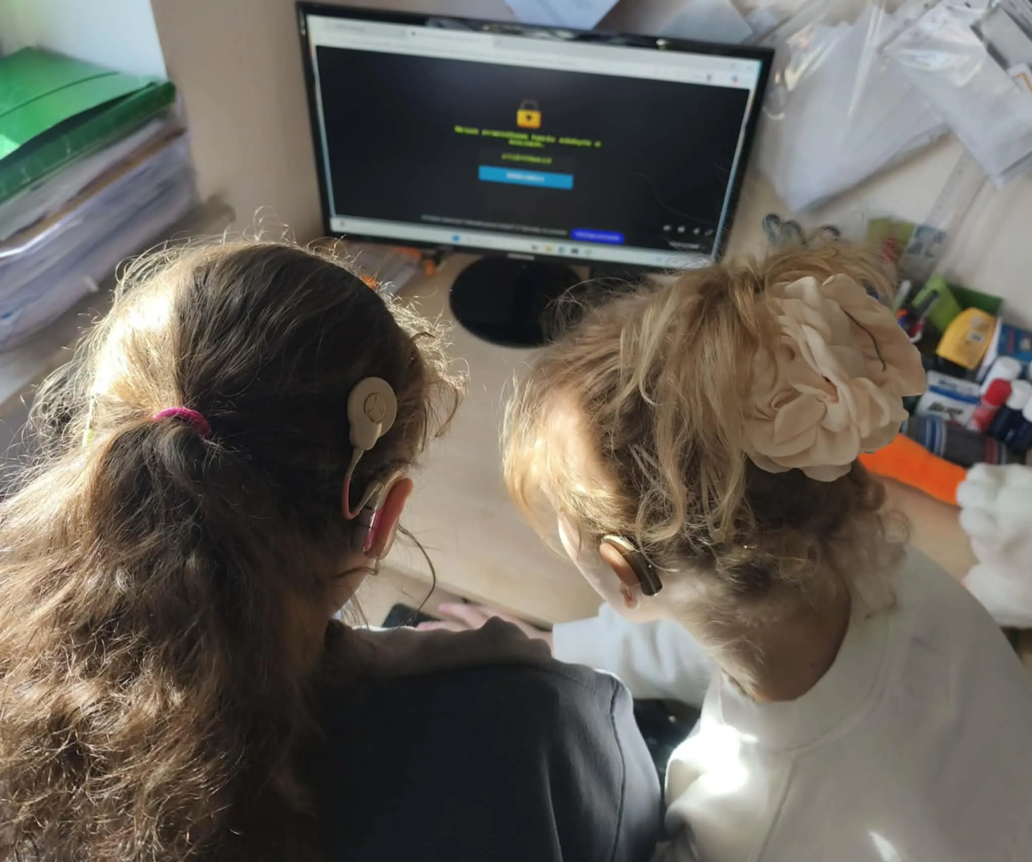 Dwie uczennice z aparatami słuchowymi pracują razem przy komputerze podczas zajęć szkolnych.