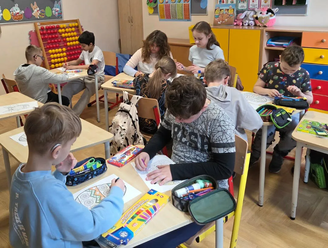 Uczniowie siedzą w klasie i kolorują wzory jajek przy szkolnych ławkach