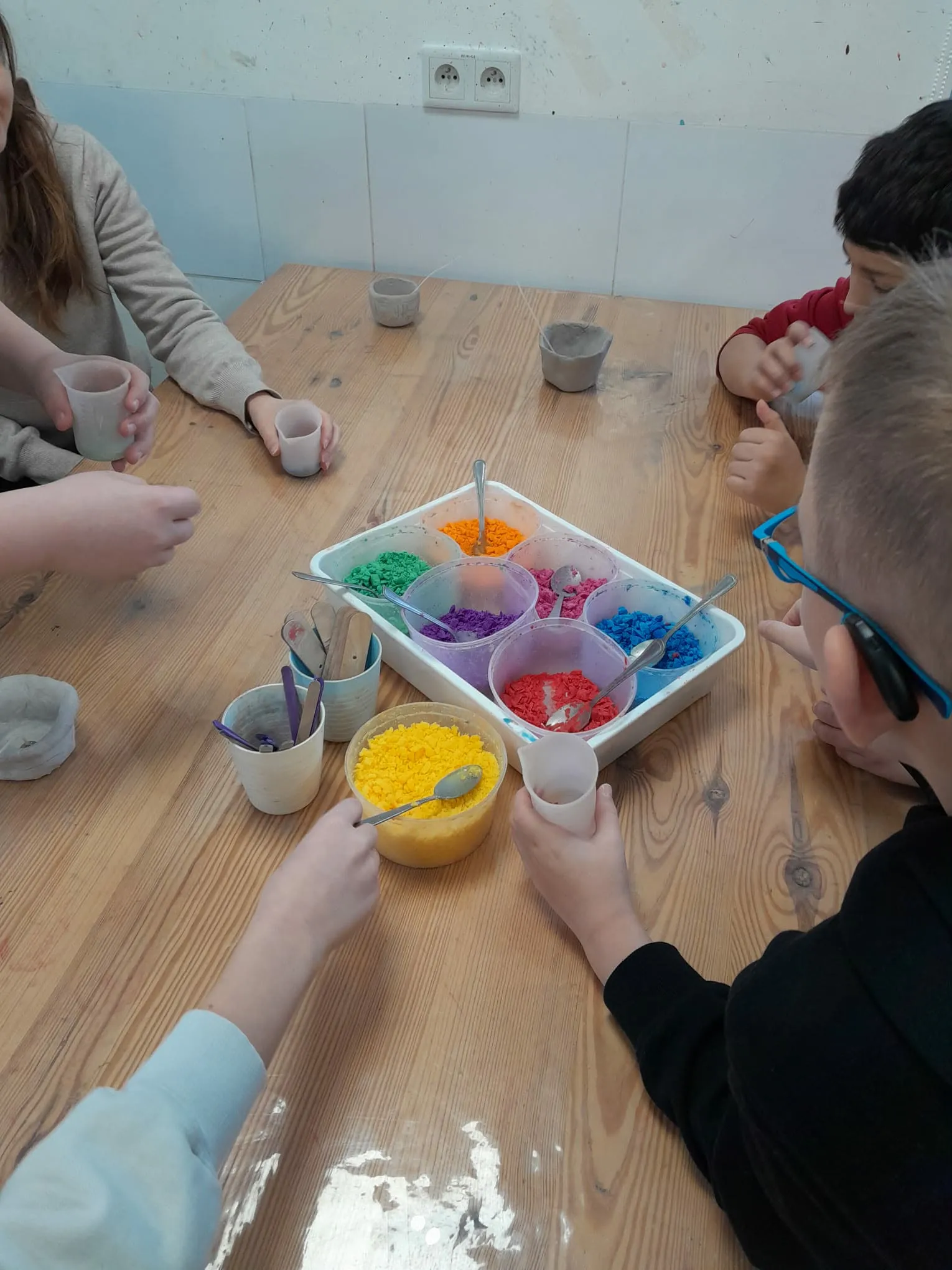 Uczniowie przy wspólnym stole wybierają kolorowy wosk z pojemników do przygotowania świec.